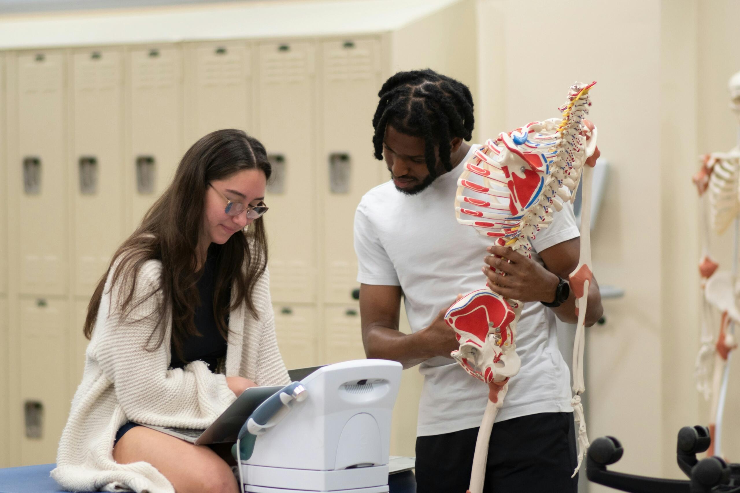 Dos alumnos estudian en el aula un modelo detallado de la columna vertebral y la pelvis humanas, junto a los casilleros y unos esqueletos anatómicos.