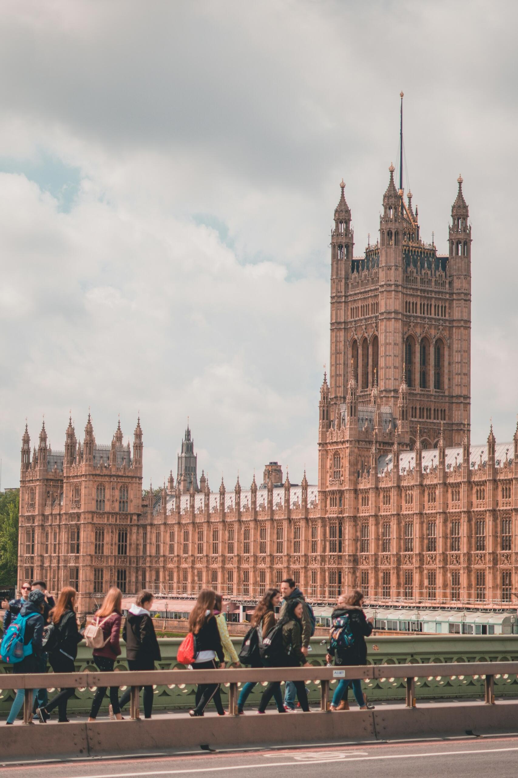 Un grupo de personas camina por un puente con la emblemática arquitectura gótica del Parlamento de Londres al fondo, bajo un cielo nublado.