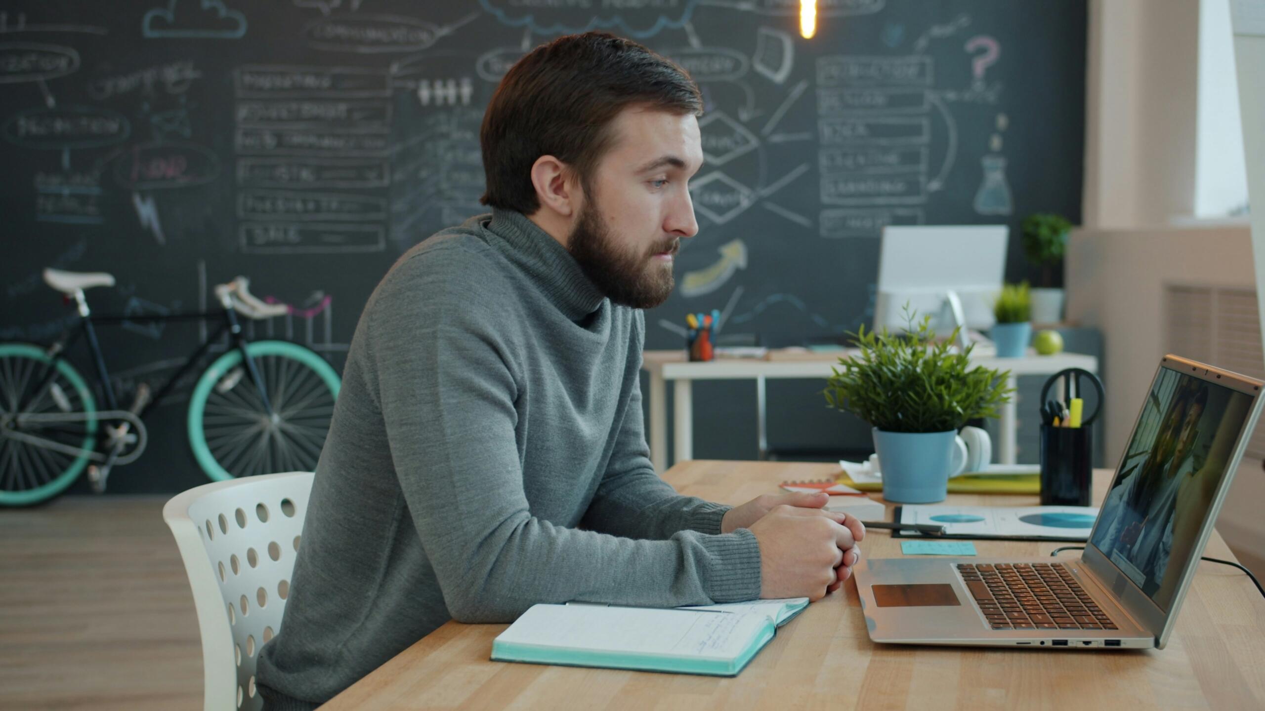 Un hombre con un jersey gris participando en una videoconferencia con su ordenador portátil en un escritorio, en una oficina creativa con una pizarra en la pared y una bicicleta al fondo.
