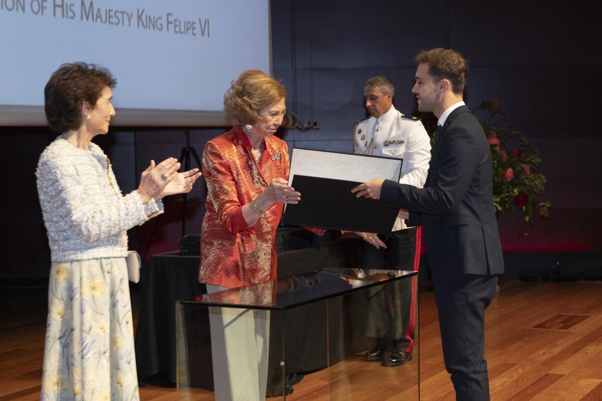 Una mujer con una chaqueta roja entrega un premio a un hombre sonriente vestido con traje en un escenario, con una mujer aplaudiendo y un hombre con un uniforme blanco formal cerca.