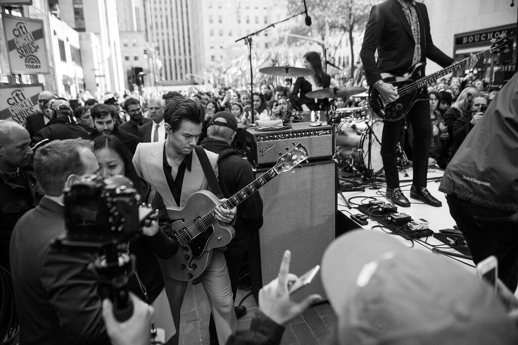 Un músico vestido con traje sostiene una guitarra, rodeado por una multitud en un concierto al aire libre, con una banda tocando al fondo.