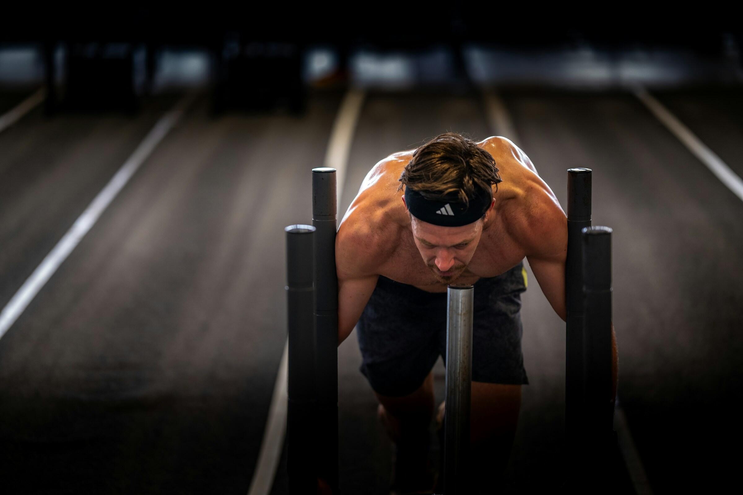 Un hombre musculoso con una cinta para el pelo empuja un trineo con pesas por una pista de gimnasio, haciendo gala de su fuerza en la parte superior del cuerpo y de su intensa concentración en el entrenamiento.