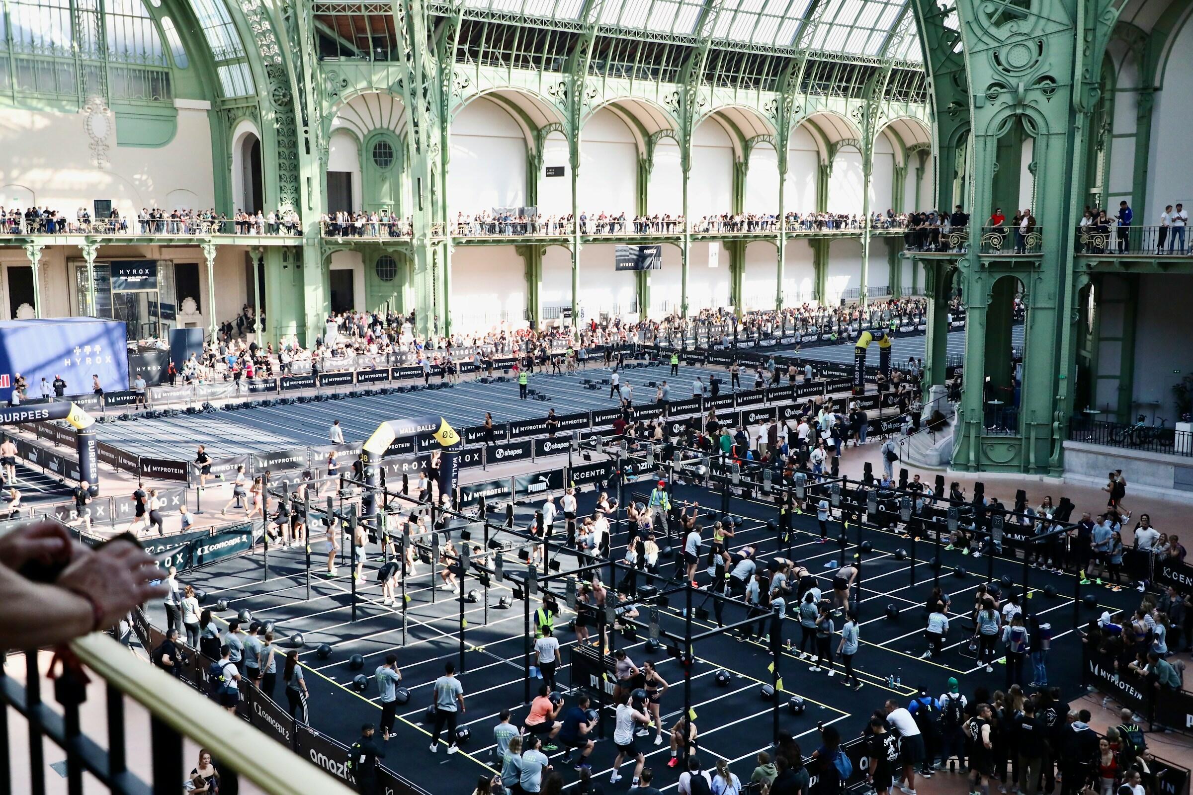 Gran evento de fitness en interior en el que numerosos participantes realizan ejercicios en estaciones de entrenamiento dentro de una gran sala abovedada con una arquitectura metálica de color verde.