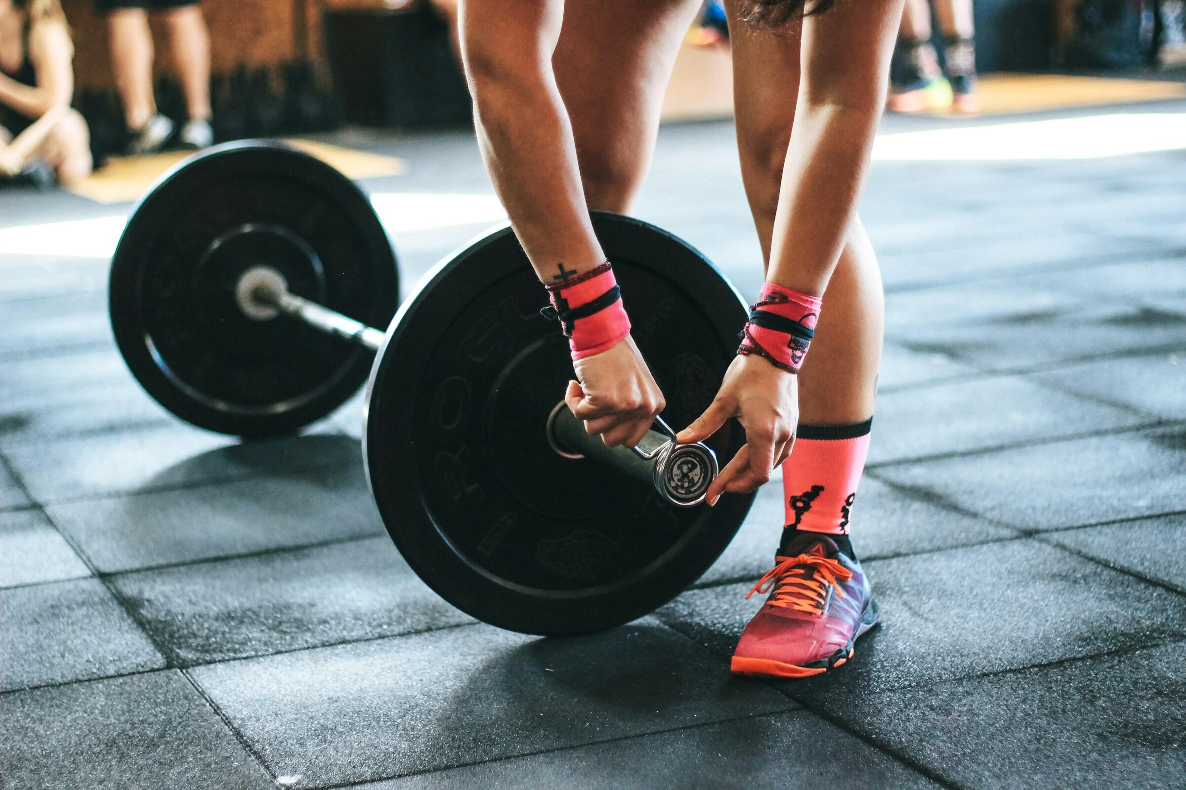 Persona colocando pesas en una barra en un gimnasio, con muñequeras rosas y zapatillas deportivas de colores.