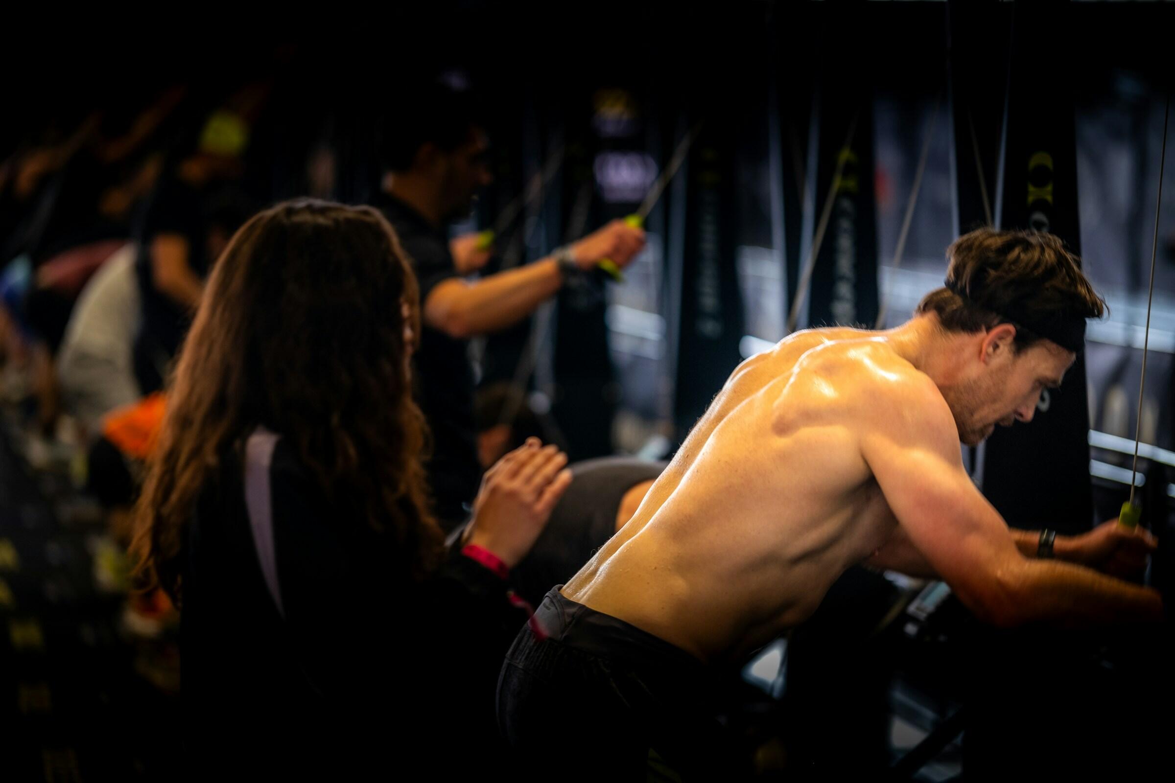 Hombre con espalda musculosa utilizando una máquina de cable en un gimnasio, con otras personas haciendo ejercicio al fondo.