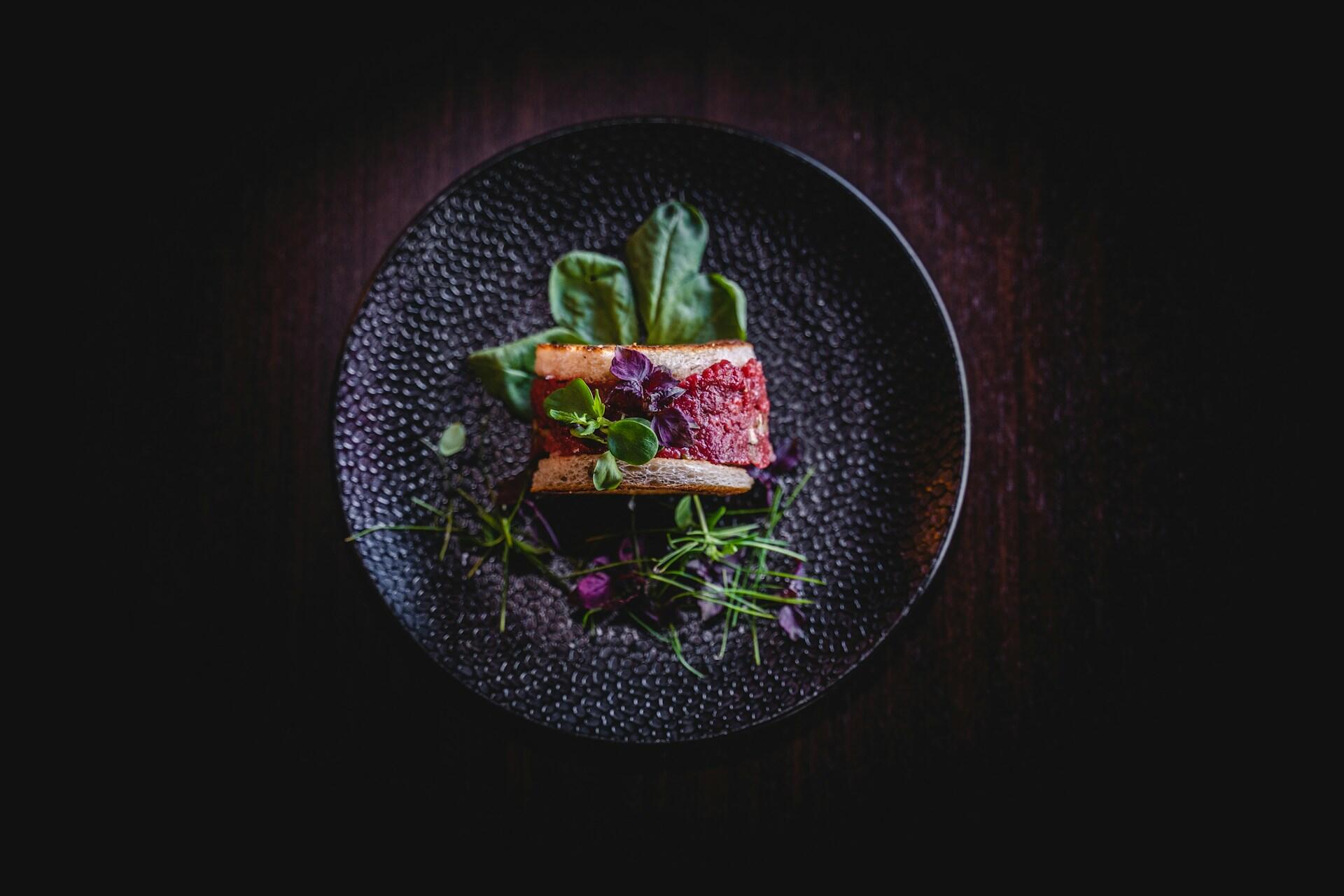 Un plato gourmet en un plato negro con textura, con un sándwich en capas de tartar de ternera, adornado con hierbas y verduras.