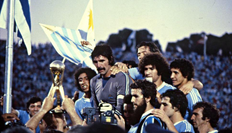 Un grupo de futbolistas radiantes, con uniformes azul claro y blancos, celebra una victoria rodeando un trofeo dorado que sostienen unas manos que vitorean. Al fondo ondean banderas.