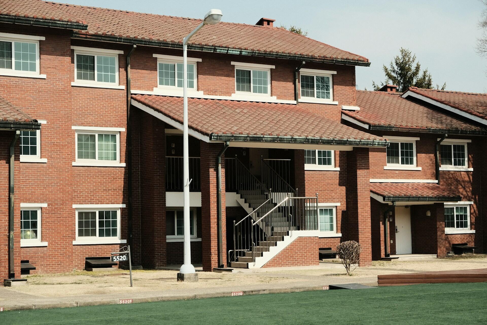 Un edificio de apartamentos de dos plantas, construido en ladrillo rojo, con una escalera central, ventanas con marcos blancos y un césped verde. La escena transmite una sensación de tranquilidad y de barrio residencial.