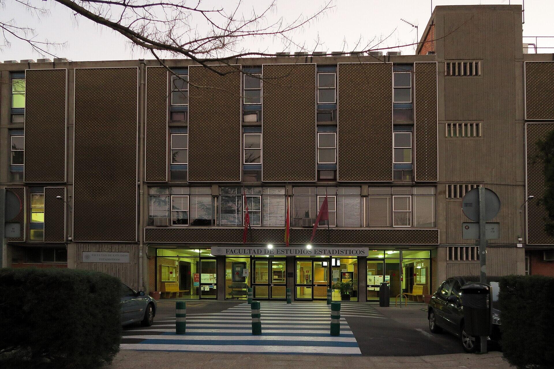 Fachada del edificio de la Facultad de Estudios Estadísticos de la universidad complutense de madrid al atardecer, con la entrada iluminada, un paso de peatones, coches aparcados y ramas de árboles sin hojas en lo alto.