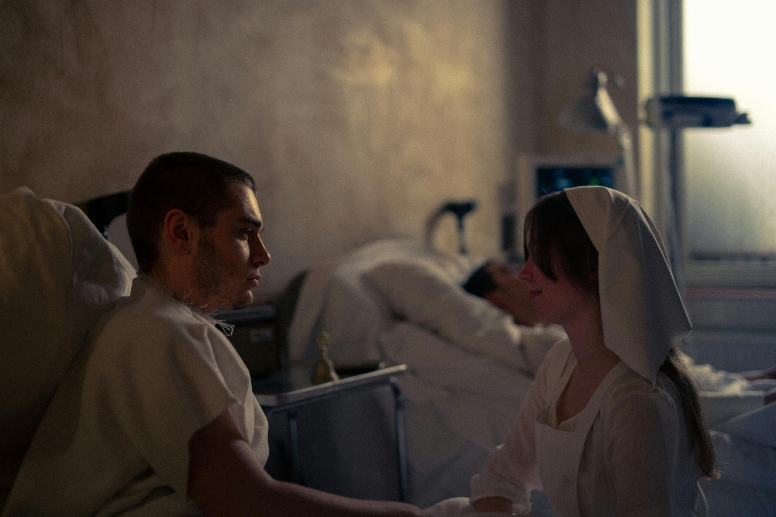 Habitación de hospital en la que una enfermera atiende a un paciente sentado en la cama, mientras otro paciente yace en el fondo, cerca de una ventana.