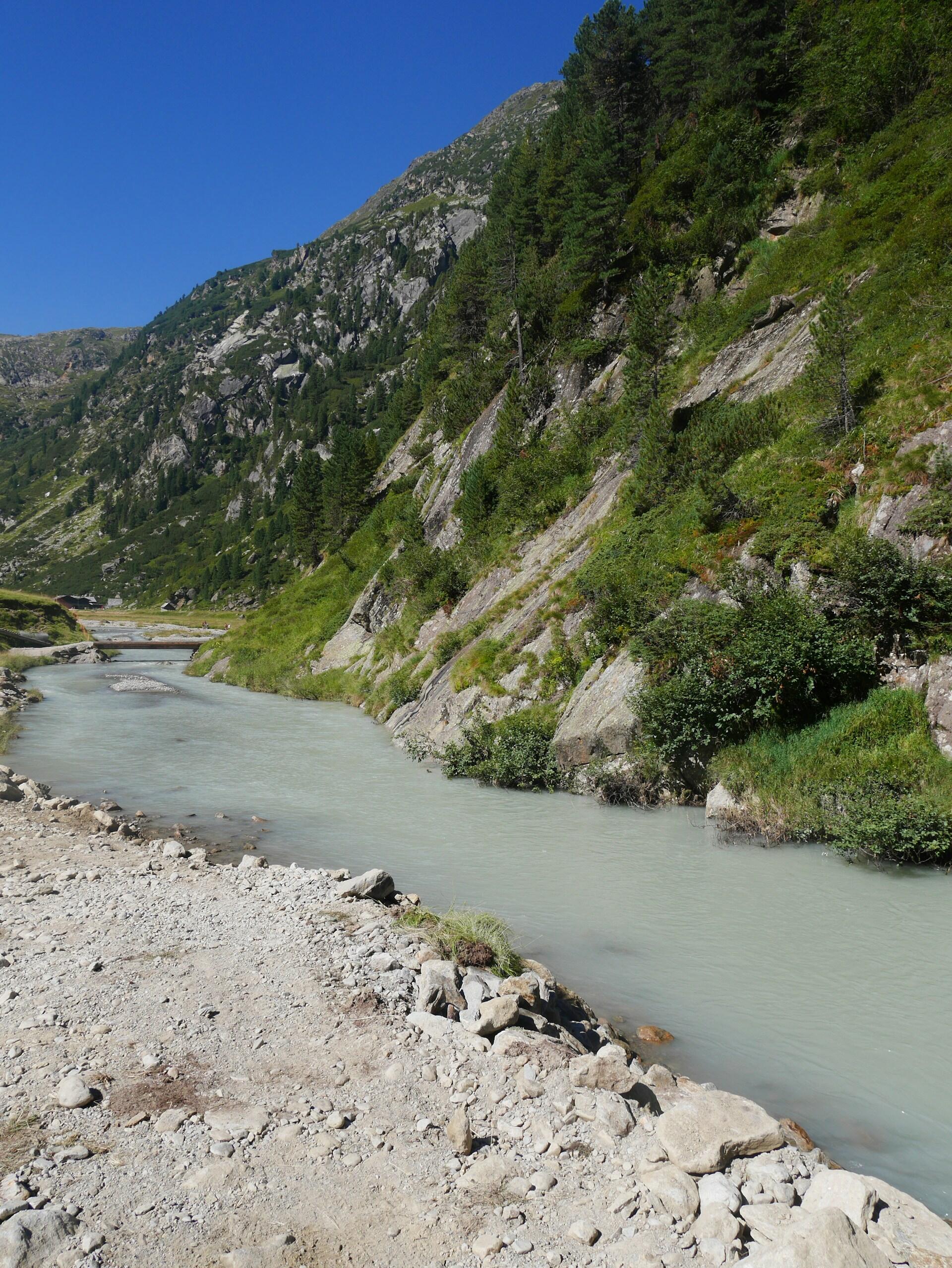 Un sereno paisaje montañoso, con un río serpenteante de aguas azul lechoso, orillas rocosas, laderas de un verde exuberante y un cielo azul claro, transmite una gran sensación de tranquilidad.
