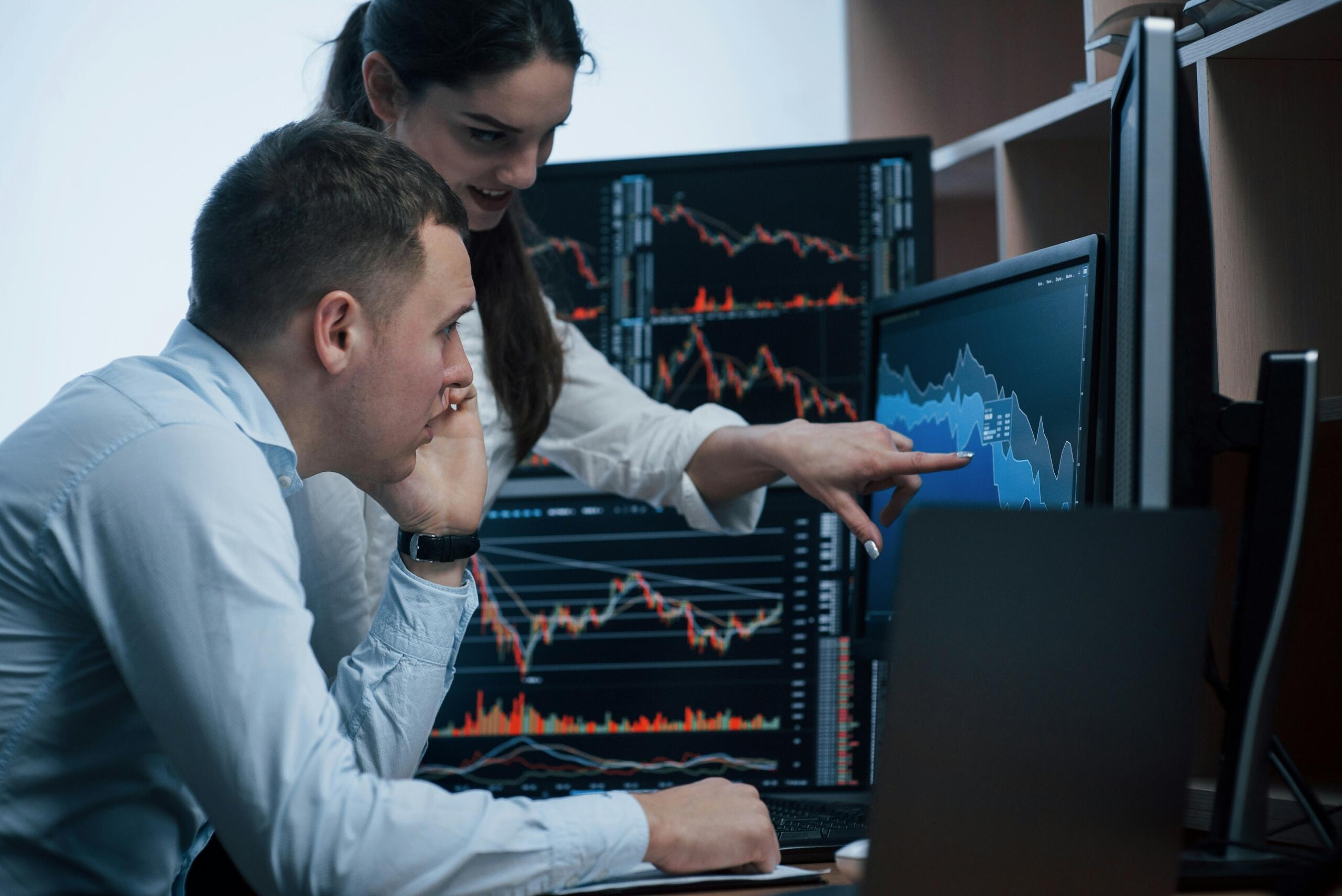 Dos profesionales analizando gráficos y datos financieros en varias pantallas de ordenador en una oficina.