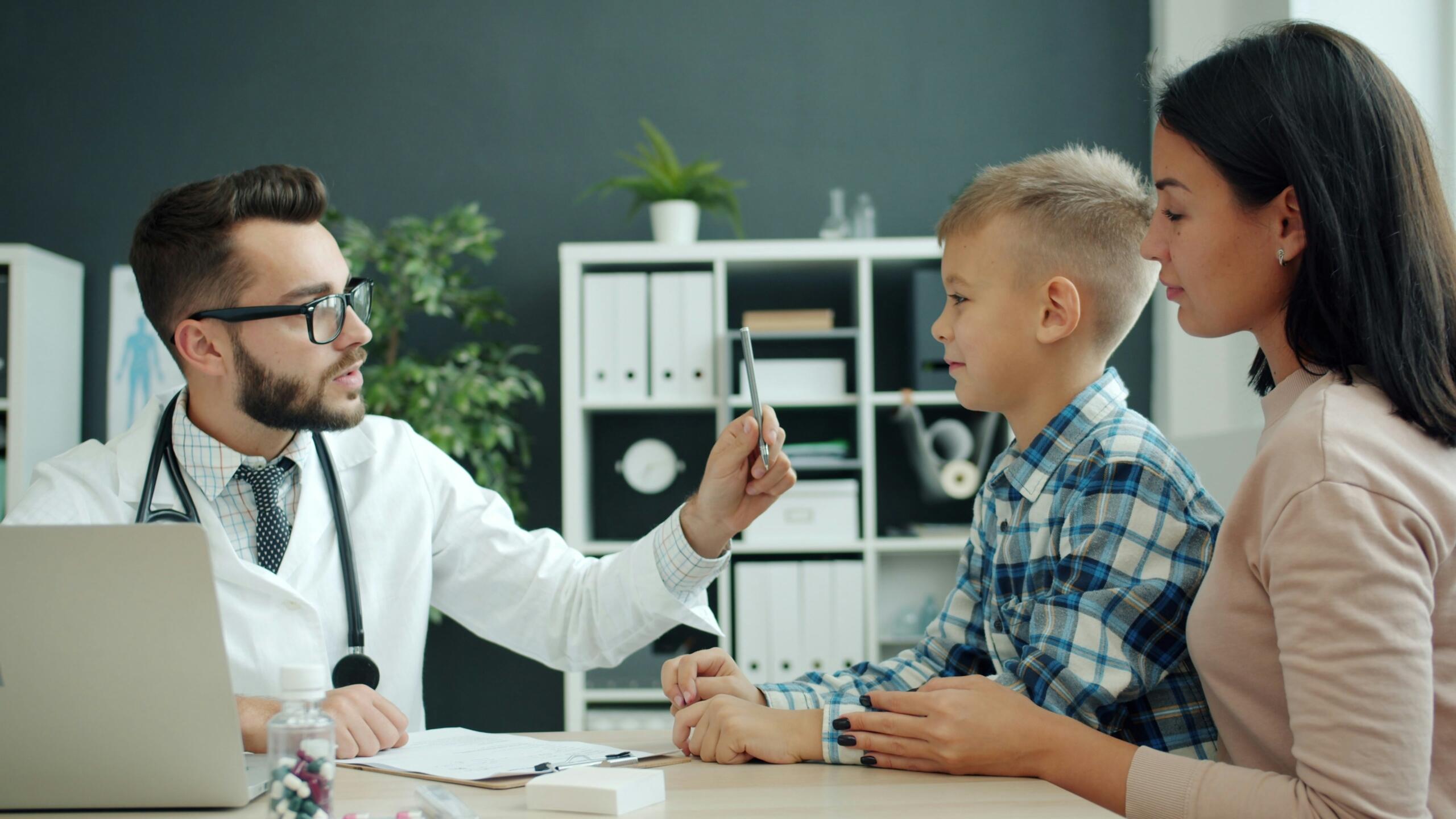 Un médico con bata blanca muestra una tableta a un niño pequeño y a su madre en una consulta. Al fondo se ven estanterías con carpetas y plantas.