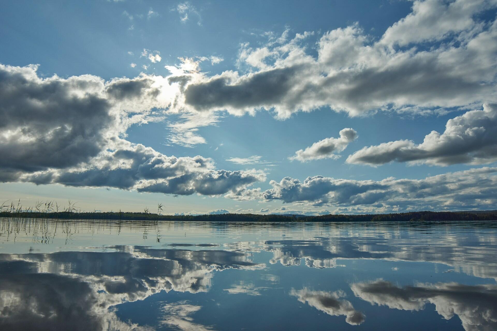 Un lago tranquilo que refleja un cielo azul parcialmente nublado, con rayos de sol que se asoman entre las nubes esponjosas, creando una atmósfera serena y apacible.
