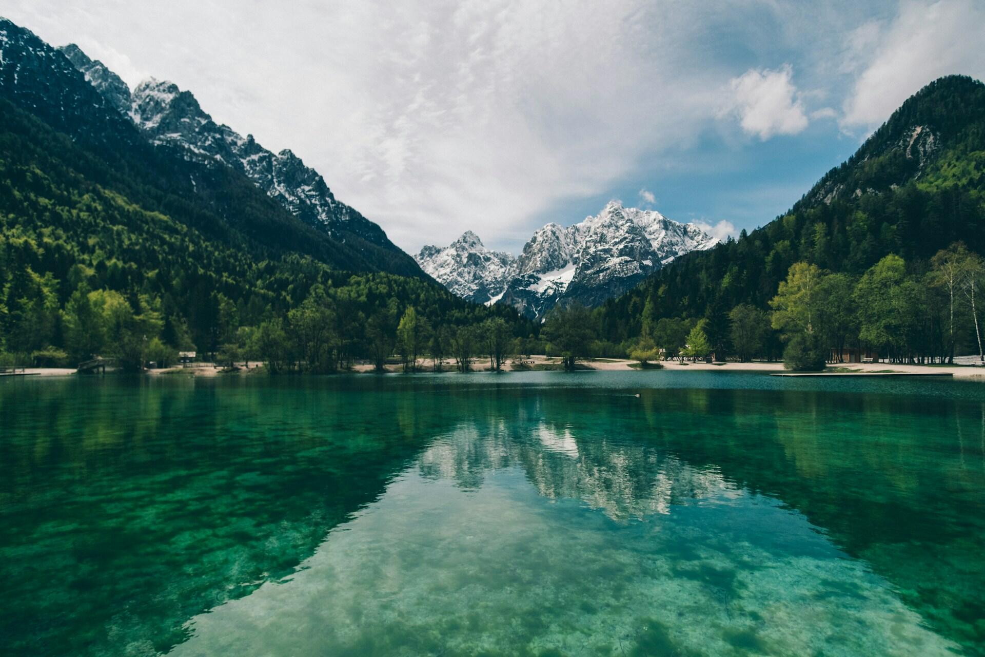 Un lago tranquilo de aguas cristalinas de color turquesa refleja las montañas nevadas y los frondosos bosques verdes bajo un cielo parcialmente nublado, lo que evoca serenidad y belleza natural.