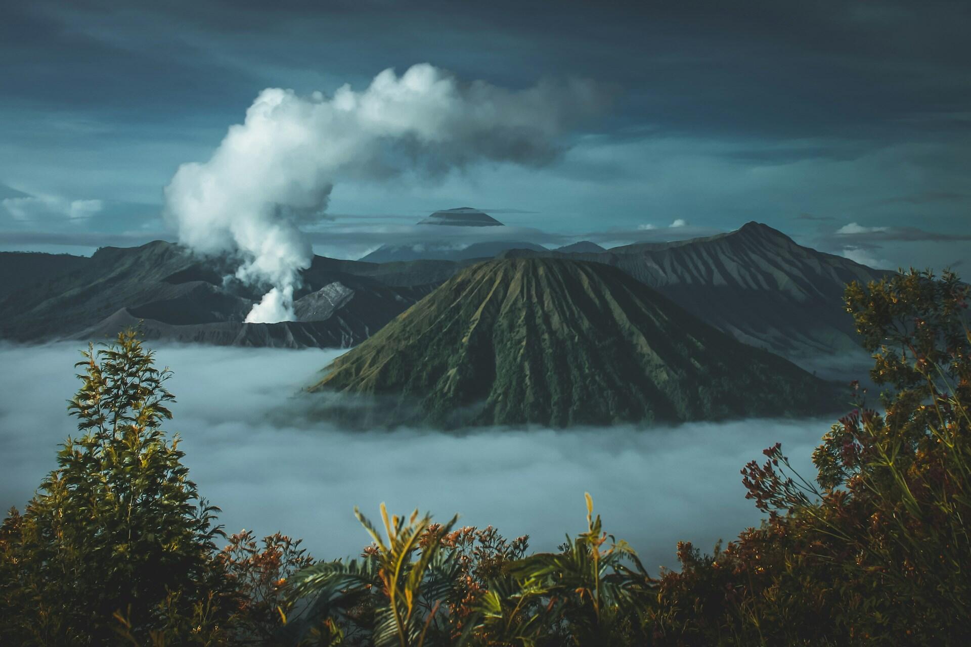 Majestuosa vista de un volcán humeante rodeado de exuberantes montañas verdes y valles envueltos en niebla, bajo un cielo cambiante y cubierto de nubes.