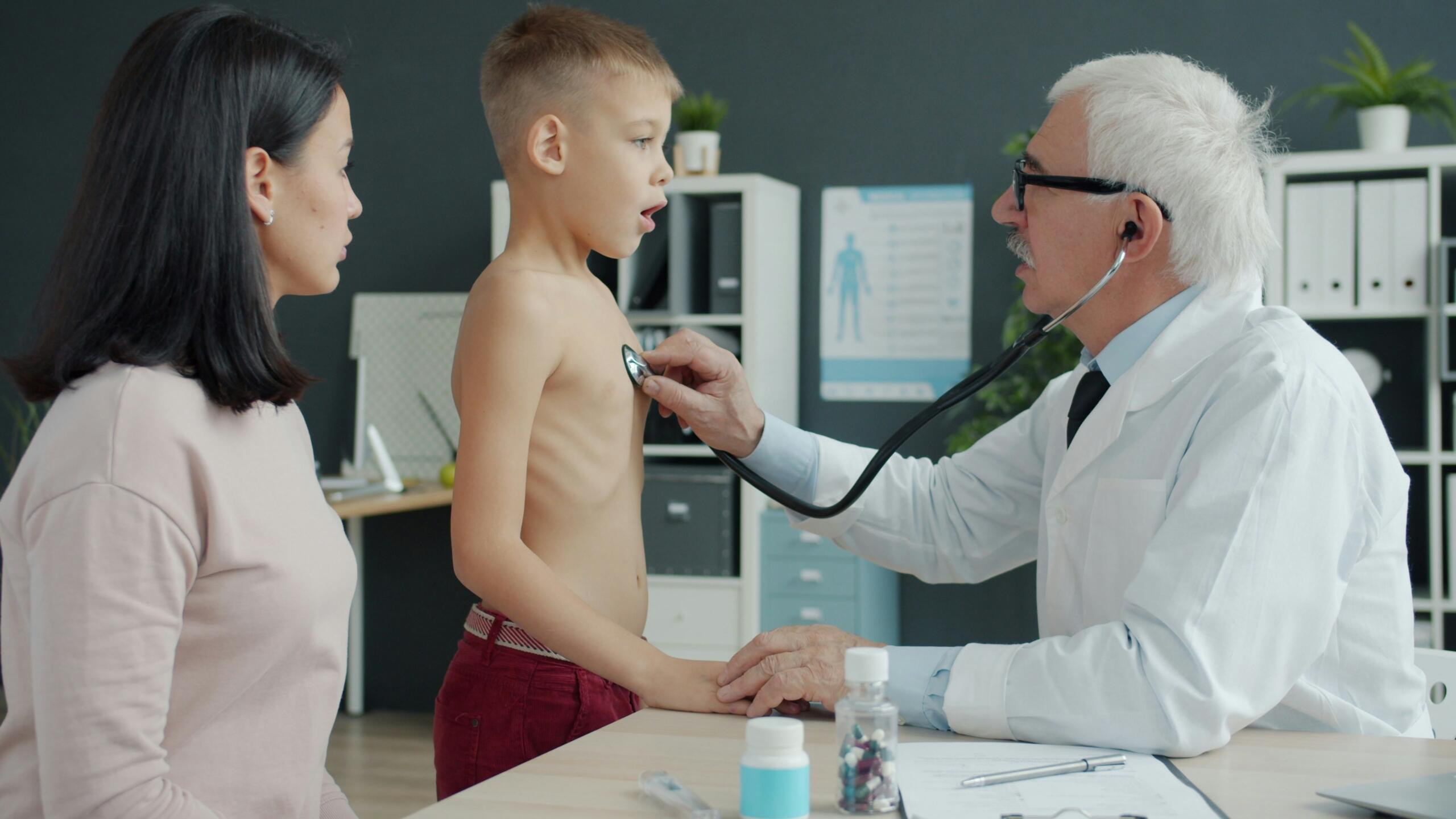 Un médico examina con un estetoscopio a un niño sin camiseta mientras le coge de la mano; una mujer observa la escena en la consulta.