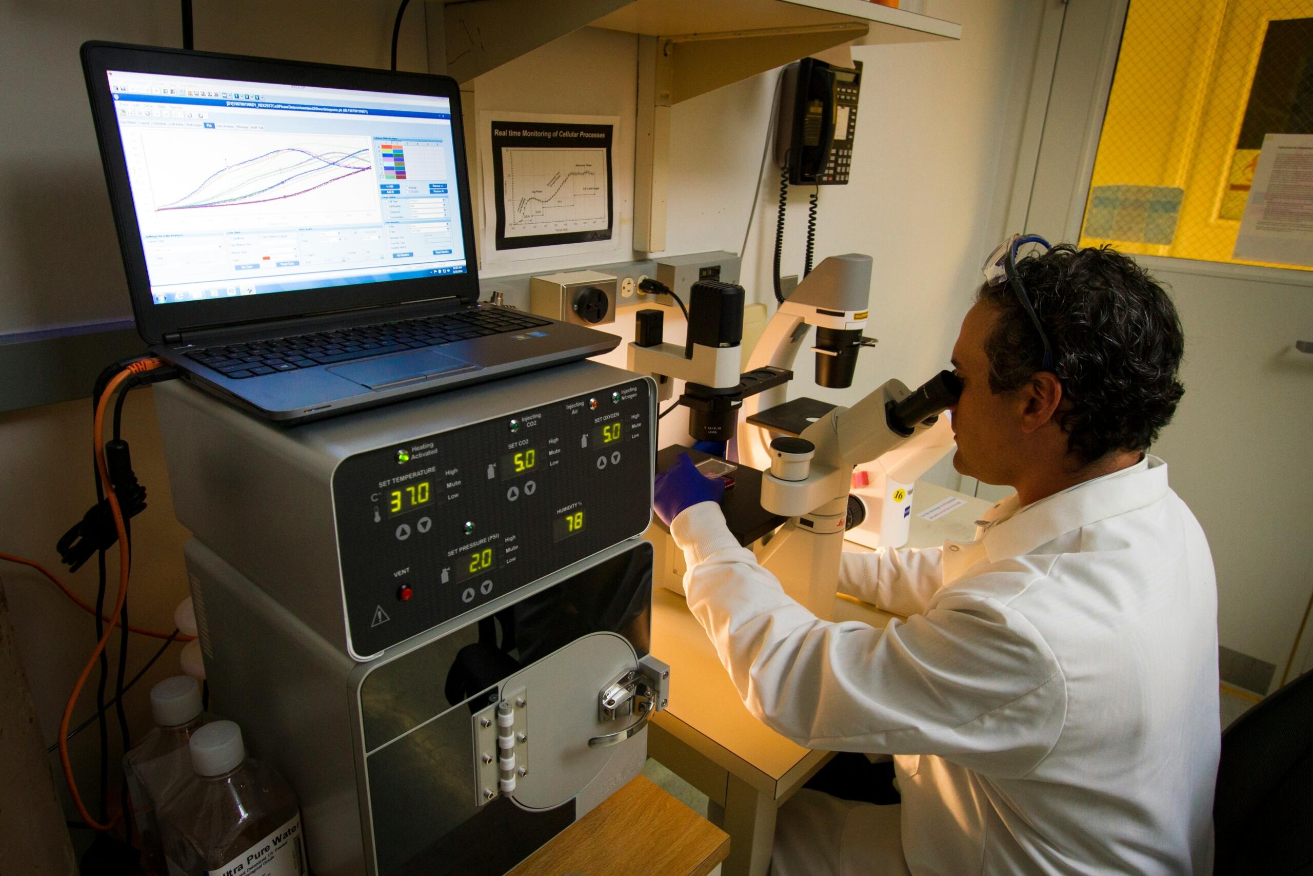 Científico con bata de laboratorio utilizando un microscopio, junto a un ordenador y equipos de monitorización, en un entorno de laboratorio.