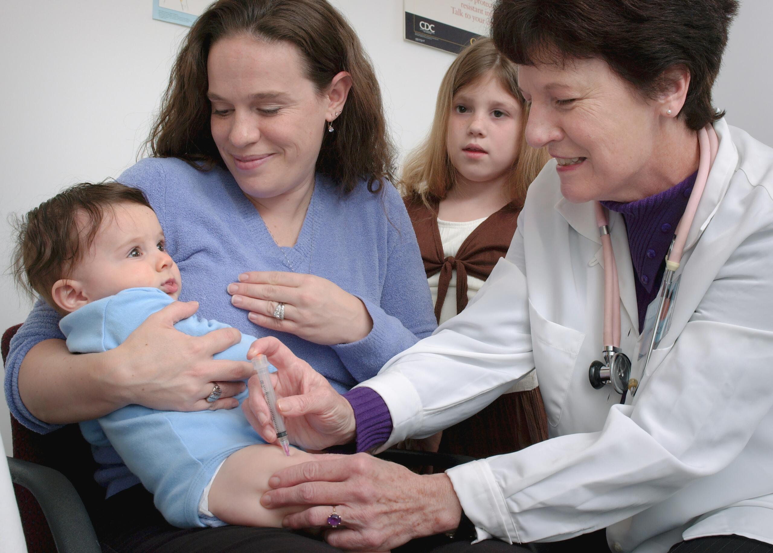 Un bebé recibe una vacuna de un médico con bata blanca mientras su madre, sonriente, lo sostiene en brazos. Una niña curiosa observa la escena al fondo, lo que transmite una atmósfera de cariño y atención.