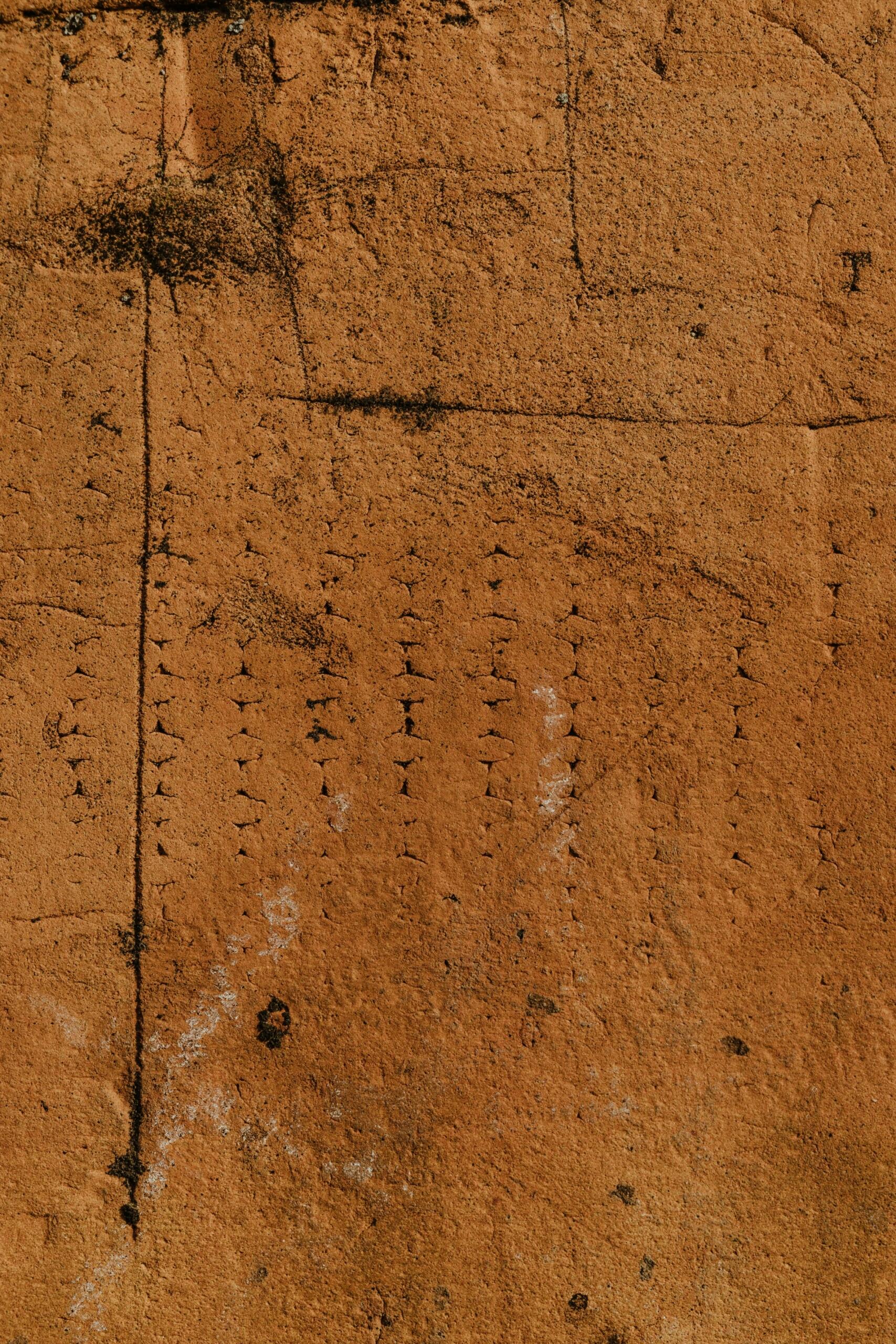 Primer plano de una superficie de textura rugosa, de color marrón anaranjado, con marcas negras verticales y horizontales y tenues manchas blancas.