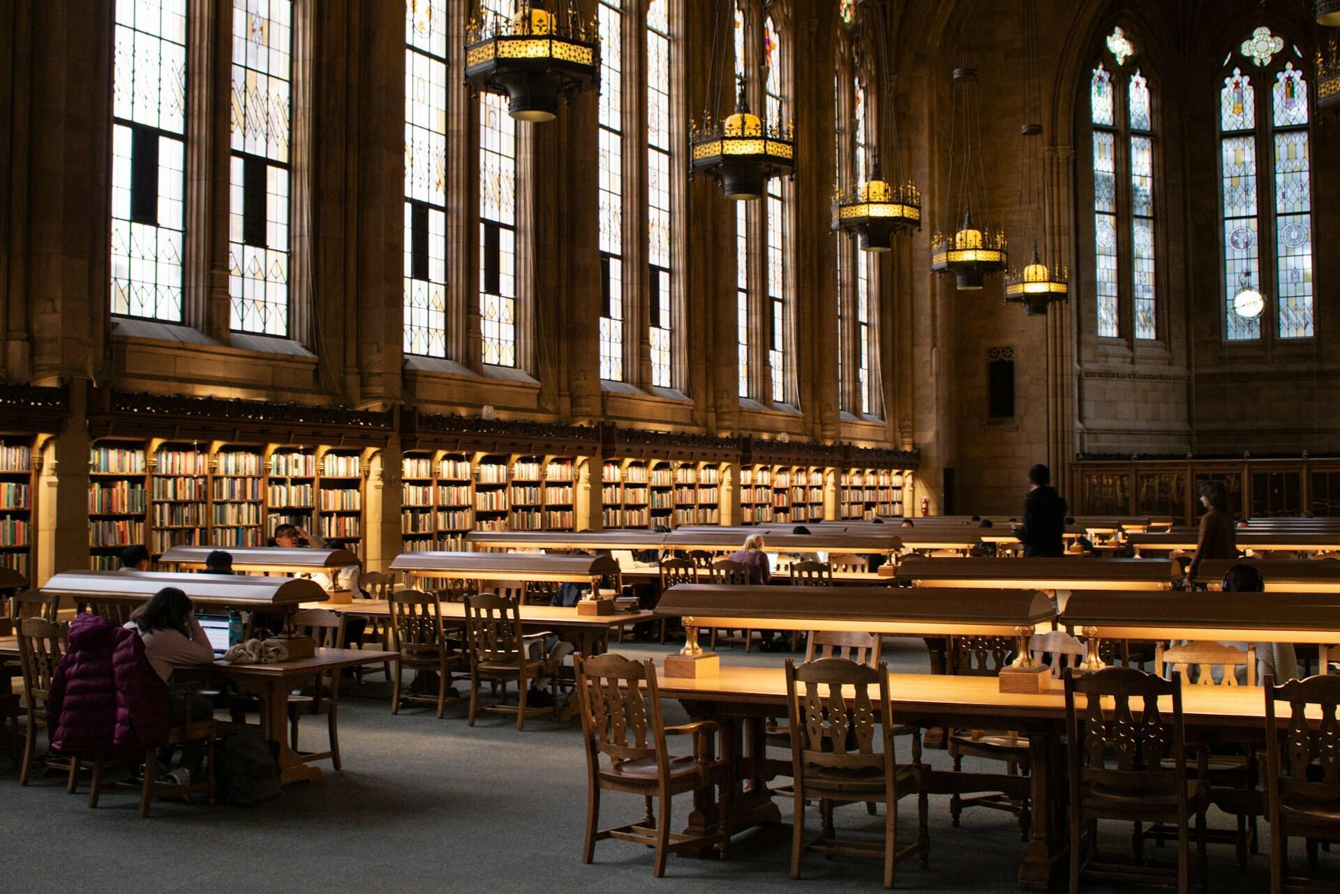 Interior de una gran biblioteca histórica con altos vitrales, mesas de madera, lámparas de lectura y gente estudiando.