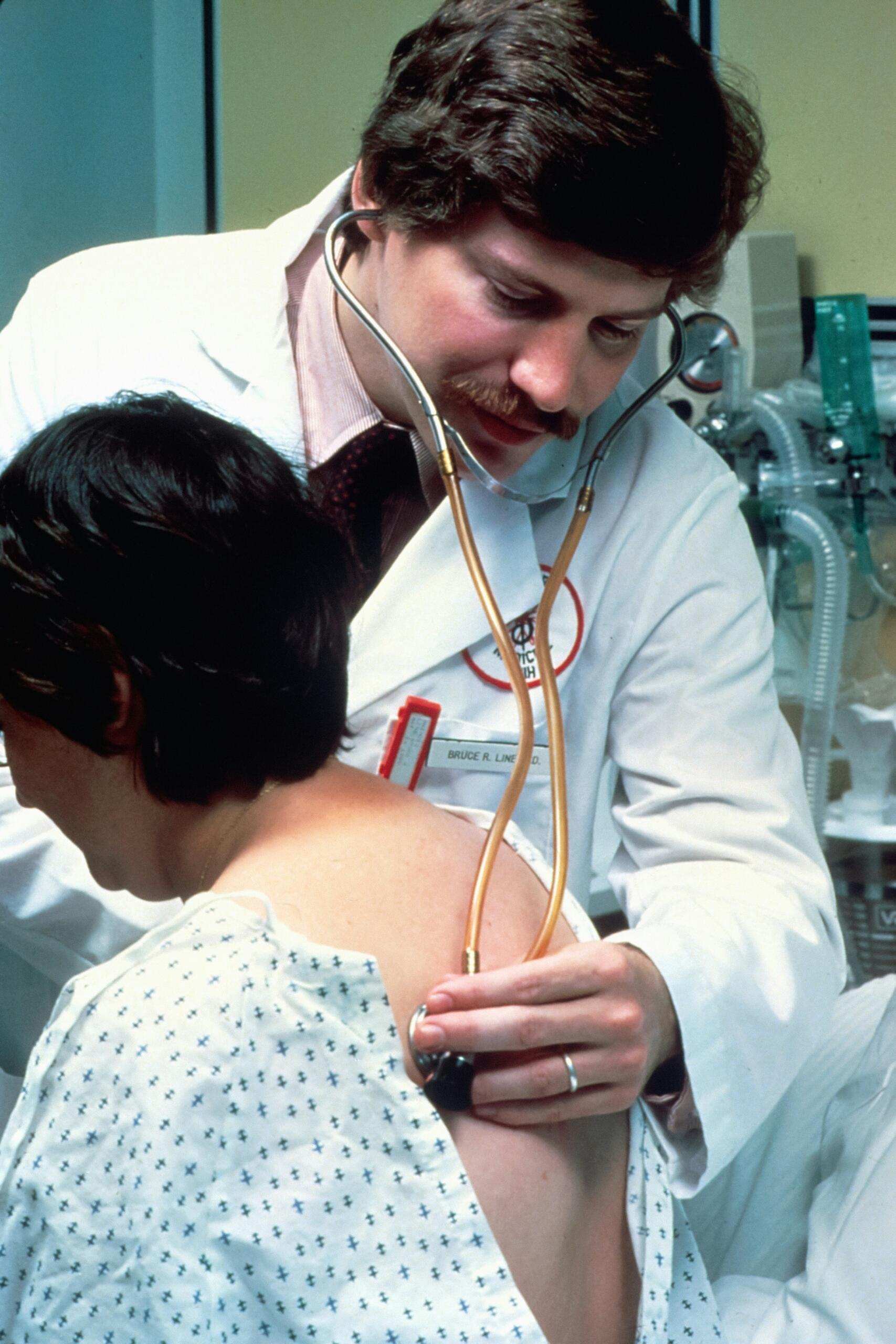 Médico con bata blanca que utiliza un estetoscopio para examinar la espalda de un paciente en un entorno clínico.