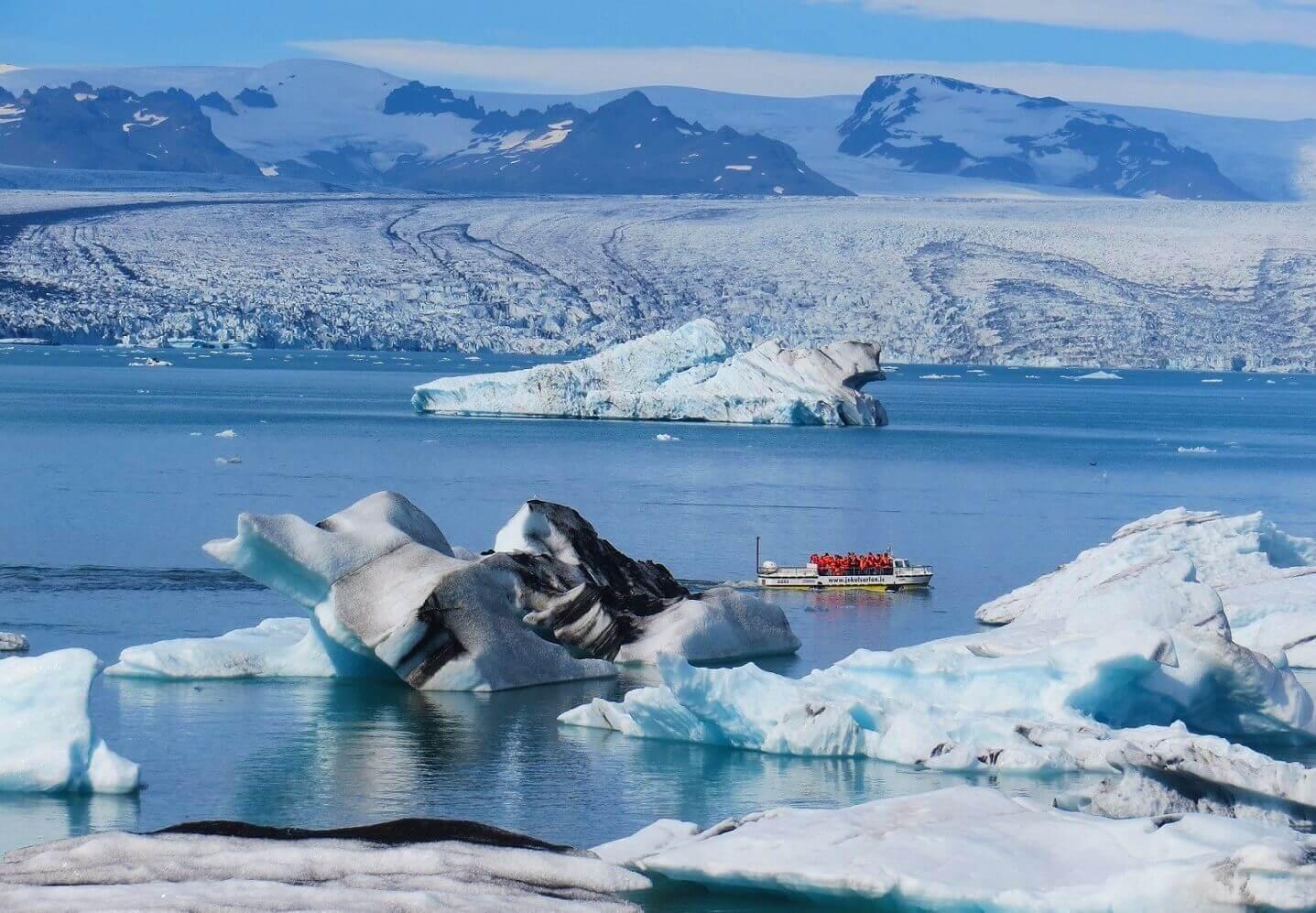 Un sereno paisaje helado con icebergs azules y blancos flotando en un lago tranquilo. En primer plano, bajo un enorme glaciar, se ve una embarcación con personas vestidas con chaquetas rojas.
