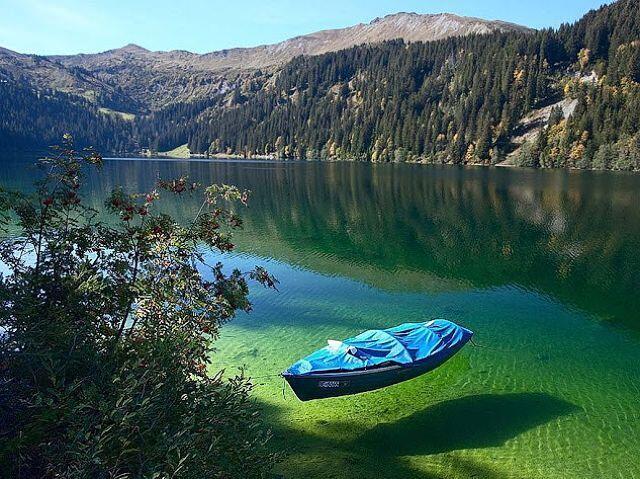 Una pequeña embarcación de cubierta azul flota en un lago verde y cristalino, rodeado de exuberantes montañas cubiertas de pinos. La escena transmite tranquilidad y belleza natural.