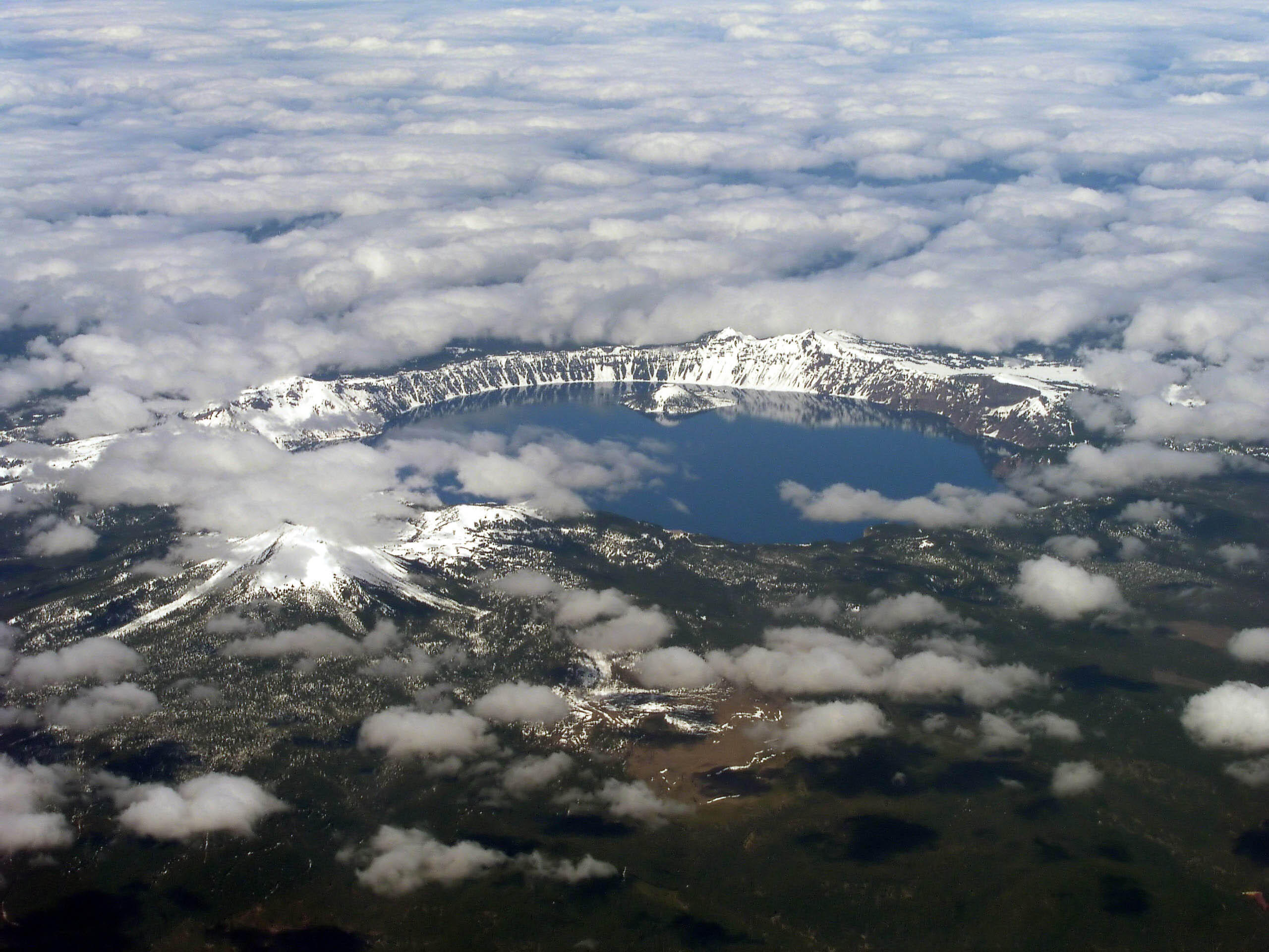 Vista aérea del lago Monte Mazama, en Oregón, rodeado por un borde nevado. Unas nubes esponjosas flotan sobre las aguas azules, creando una escena serena y majestuosa.