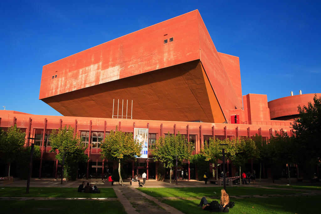 El moderno edificio de l campus de leganes de la universidad uc3m de ladrillo rojo con una gran sección superior en voladizo, árboles y gente descansando sobre el césped bajo un cielo azul y despejado.