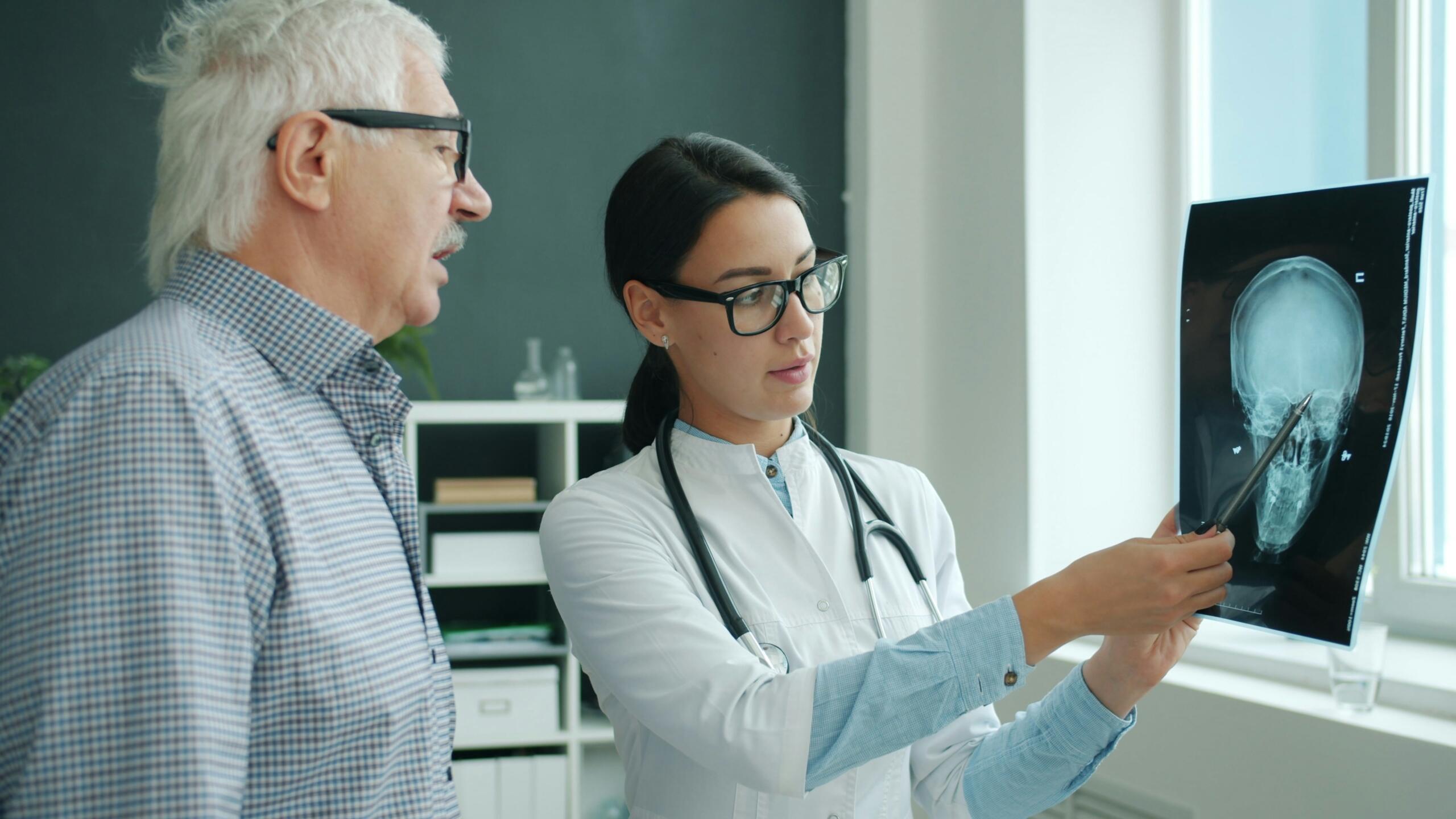 Una doctora con gafas le muestra a un anciano una radiografía del cerebro en una consulta luminosa. Ella señala la imagen mientras le da explicaciones. La escena transmite atención y cariño.