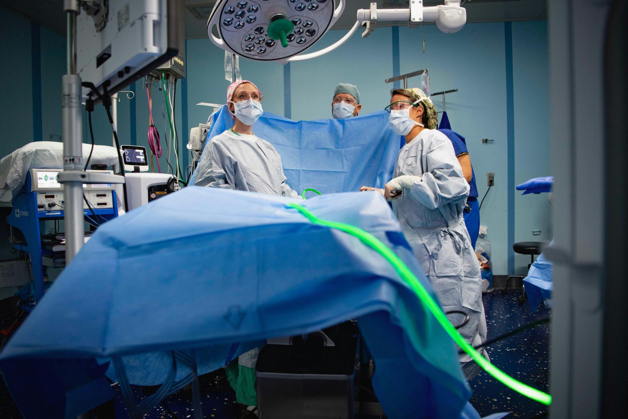 Equipo quirúrgico con batas protectoras y mascarillas preparándose para una intervención en un quirófano equipado con material médico y luces de techo.
