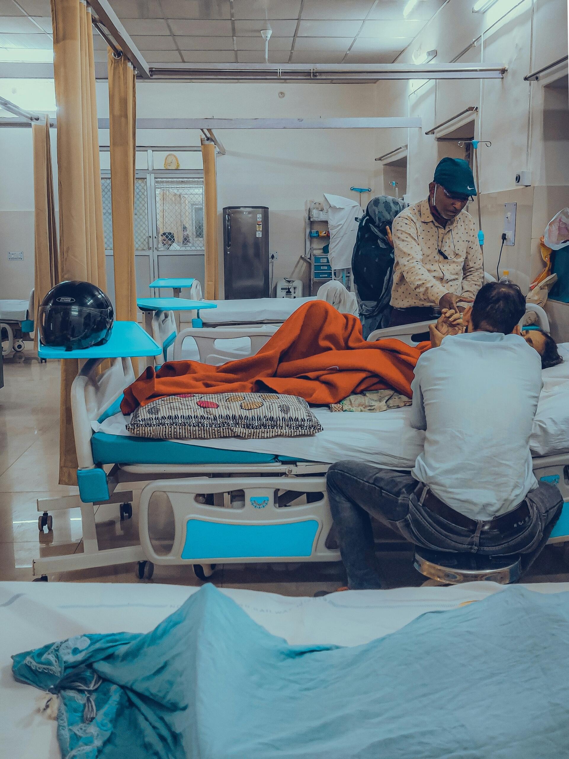 Dos personas atienden a un paciente que yace en una cama de hospital, cubierto con mantas, en una habitación de hospital en la que se ven equipos médicos y otras camas.