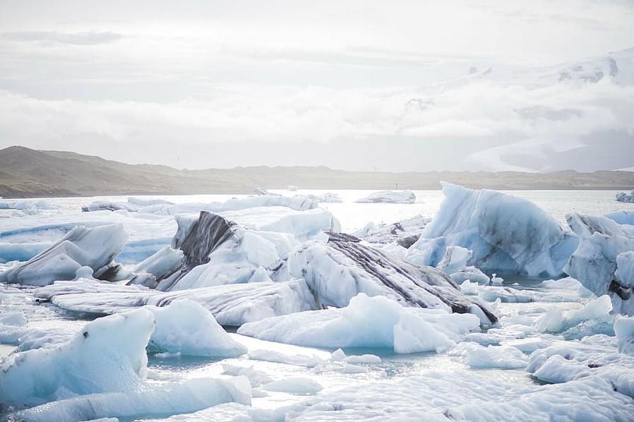 Los icebergs flotan en una serena laguna de color azul hielo bajo un cielo nublado. Al fondo se divisan montañas escarpadas, creando una escena tranquila y gélida.