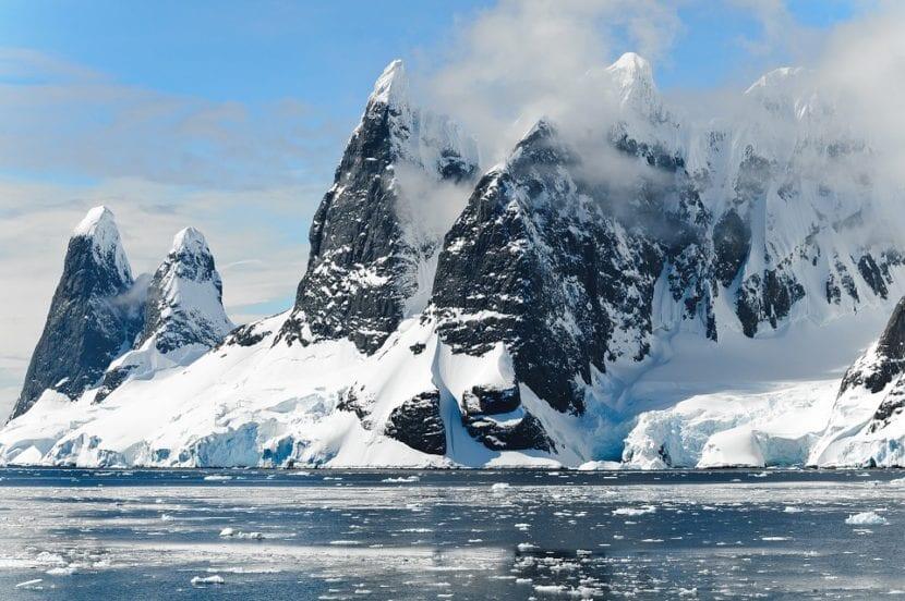 Imponentes picos nevados se alzan majestuosos sobre las aguas heladas en un escarpado paisaje antártico. Nubes bajas se desplazan alrededor de las oscuras laderas de las montañas.