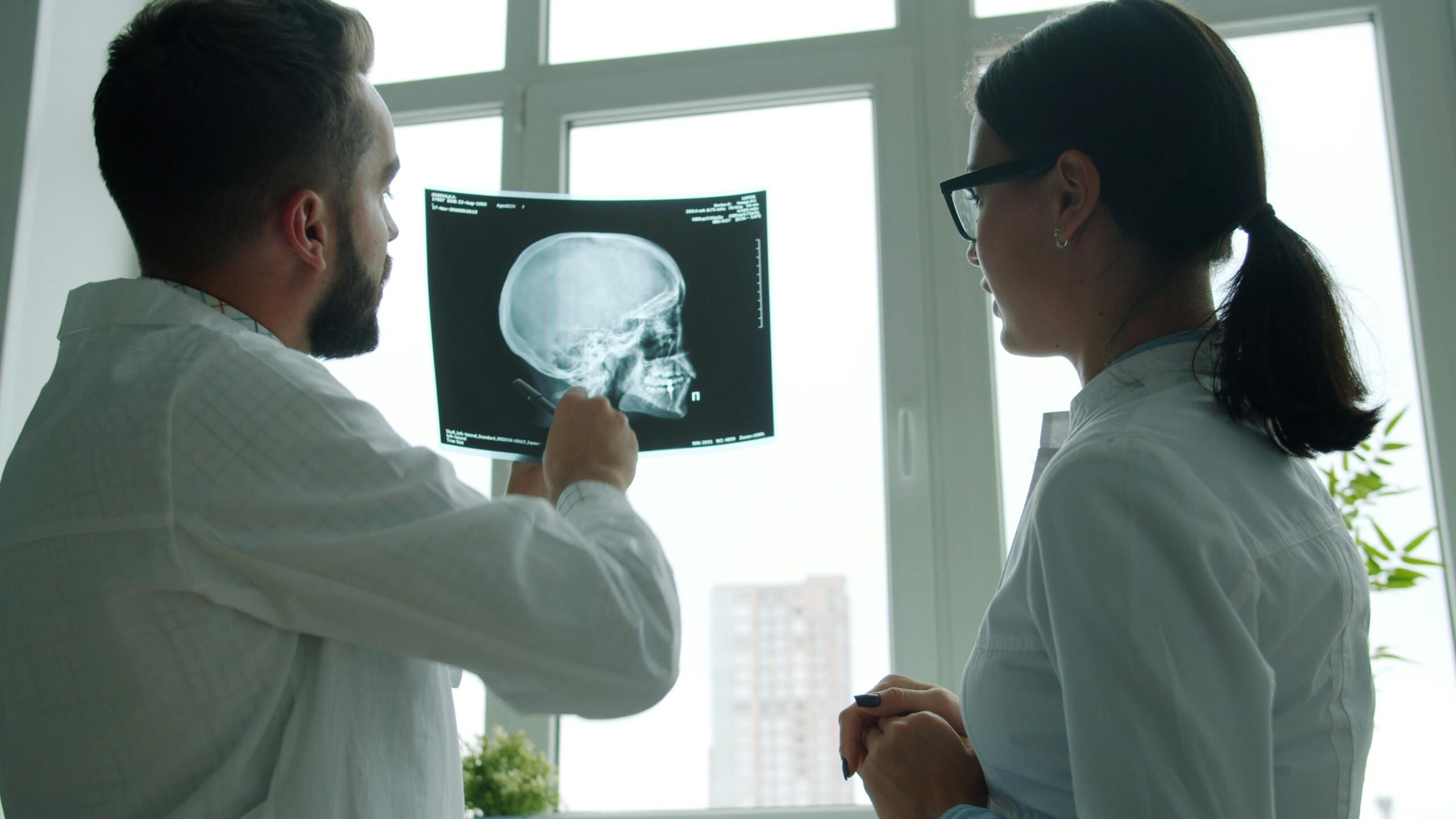 Dos médicos con bata blanca examinan una radiografía de cráneo junto a una ventana en una consulta médica.