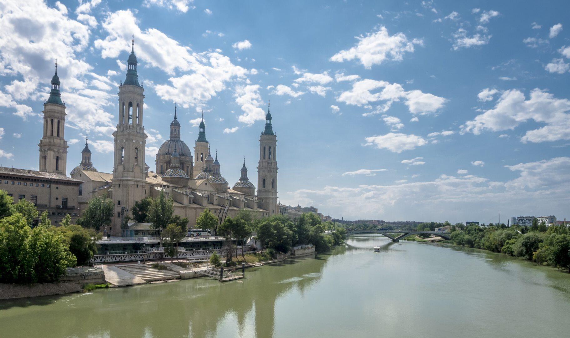 Una majestuosa catedral con múltiples agujas se alza junto a un tranquilo río bajo un cielo parcialmente nublado. Una exuberante vegetación bordea la orilla del río, lo que le confiere un ambiente sereno.