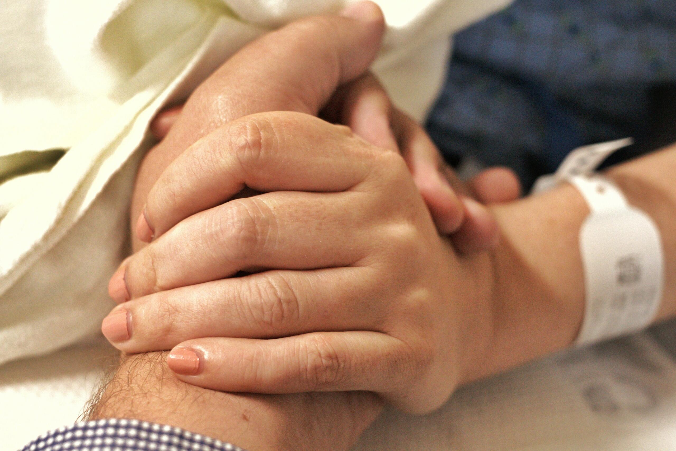 Una persona que lleva una pulsera de hospital está cogida de la mano de otra persona, brindándole consuelo y apoyo en el entorno hospitalario.
