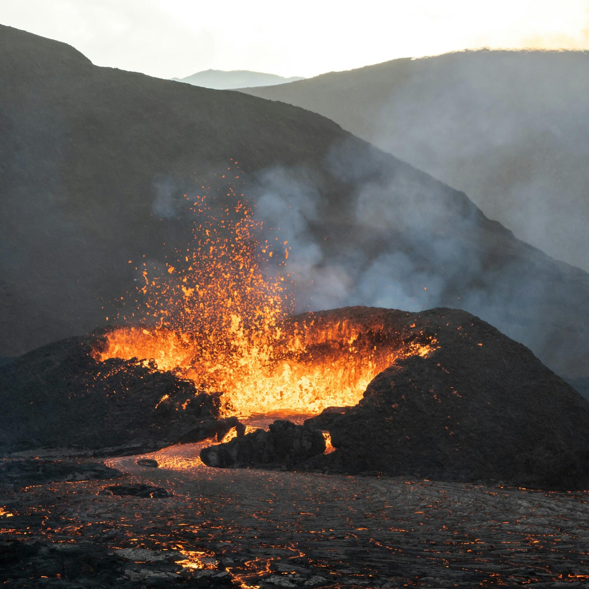 La lava ardiente brota de un volcán, y la roca fundida, de un color naranja y rojo resplandeciente, contrasta con las colinas oscuras y cubiertas de humo bajo un cielo pálido.