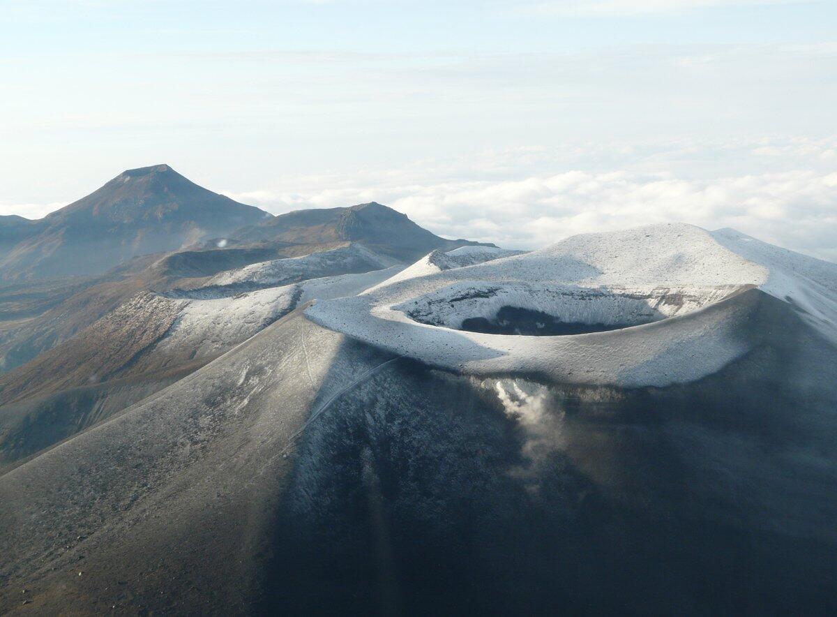 Vista aérea de cráteres volcánicos cubiertos de nieve, rodeados de montañas escarpadas y nubes. La escena transmite un paisaje natural majestuoso y sereno.