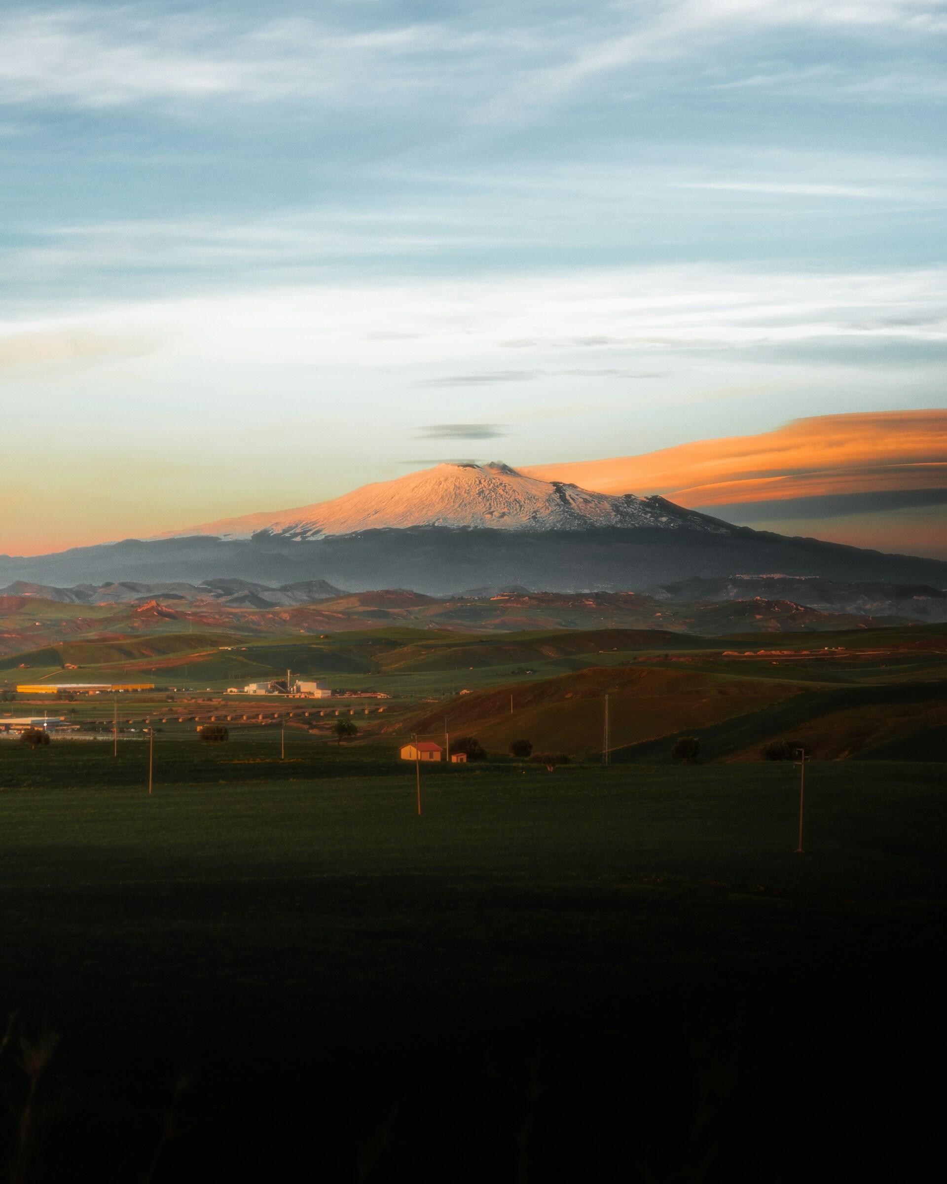 Una montaña nevada bajo un cielo de atardecer de colores vivos, con colinas verdes onduladas y edificios dispersos a sus pies, creando un paisaje tranquilo y sereno.