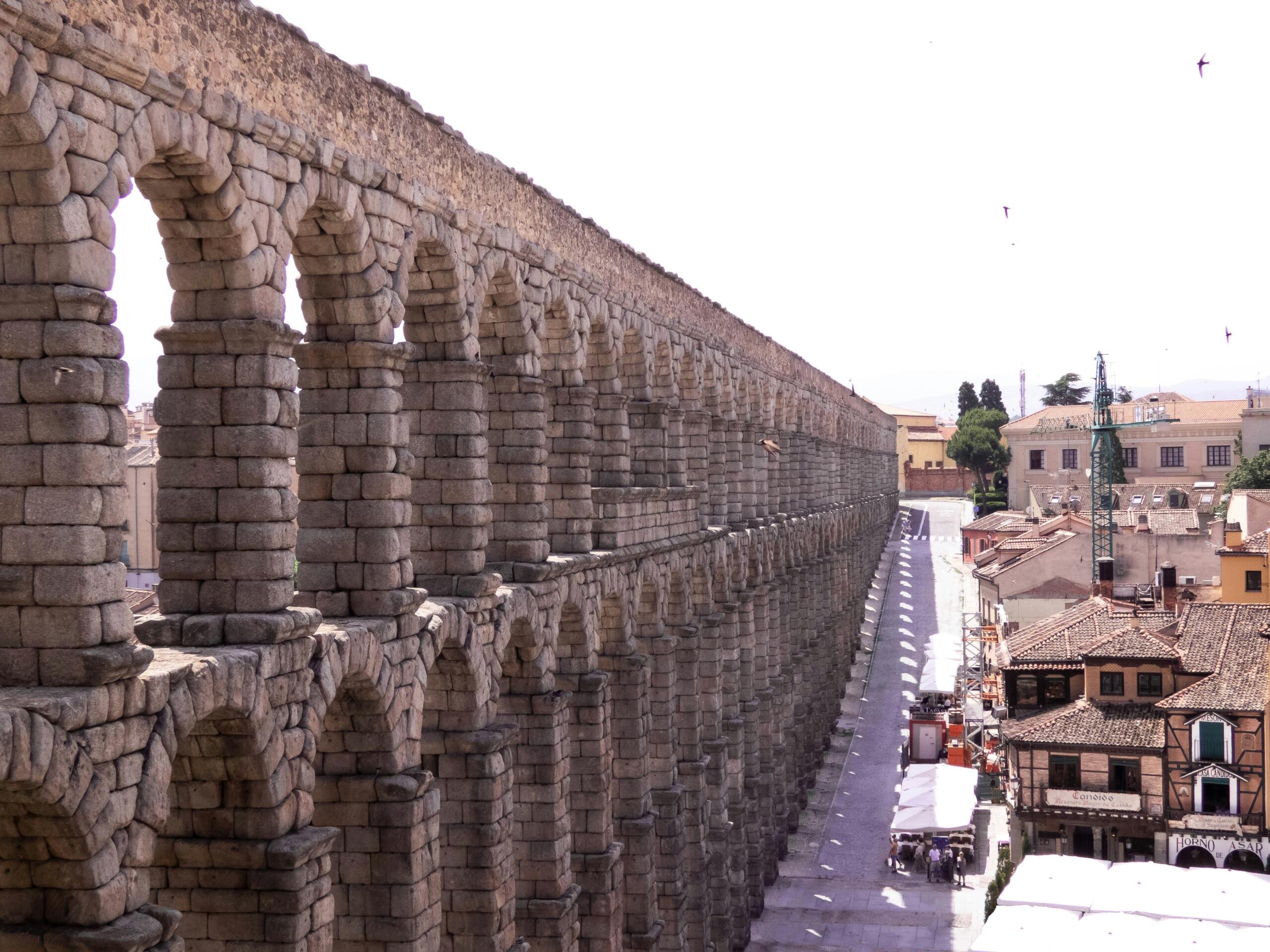 Un antiguo acueducto romano de piedra con dos hileras de arcos junto a una calle bordeada de edificios tradicionales bajo un cielo despejado.
