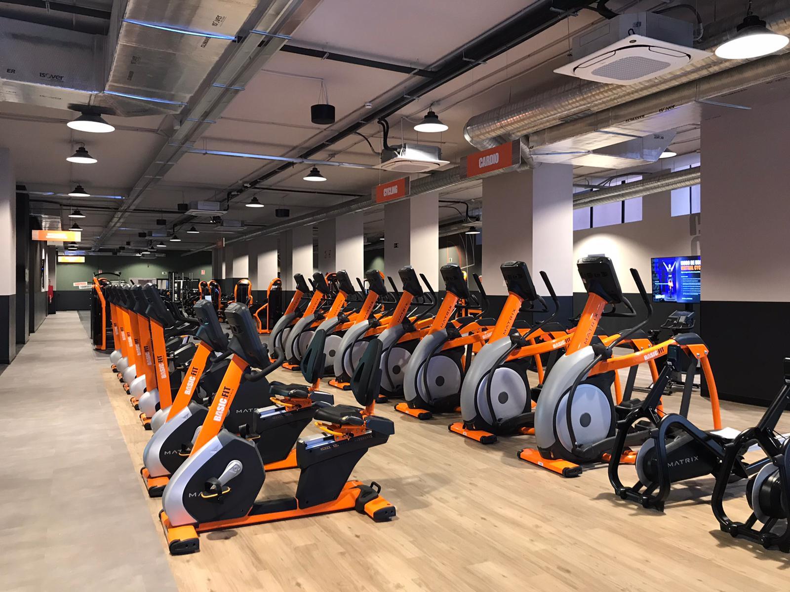 Un gimnasio moderno cuenta con filas de bicicletas estáticas y máquinas elípticas de color naranja y gris sobre un suelo de madera. El espacio está bien iluminado, es ordenado y espacioso.