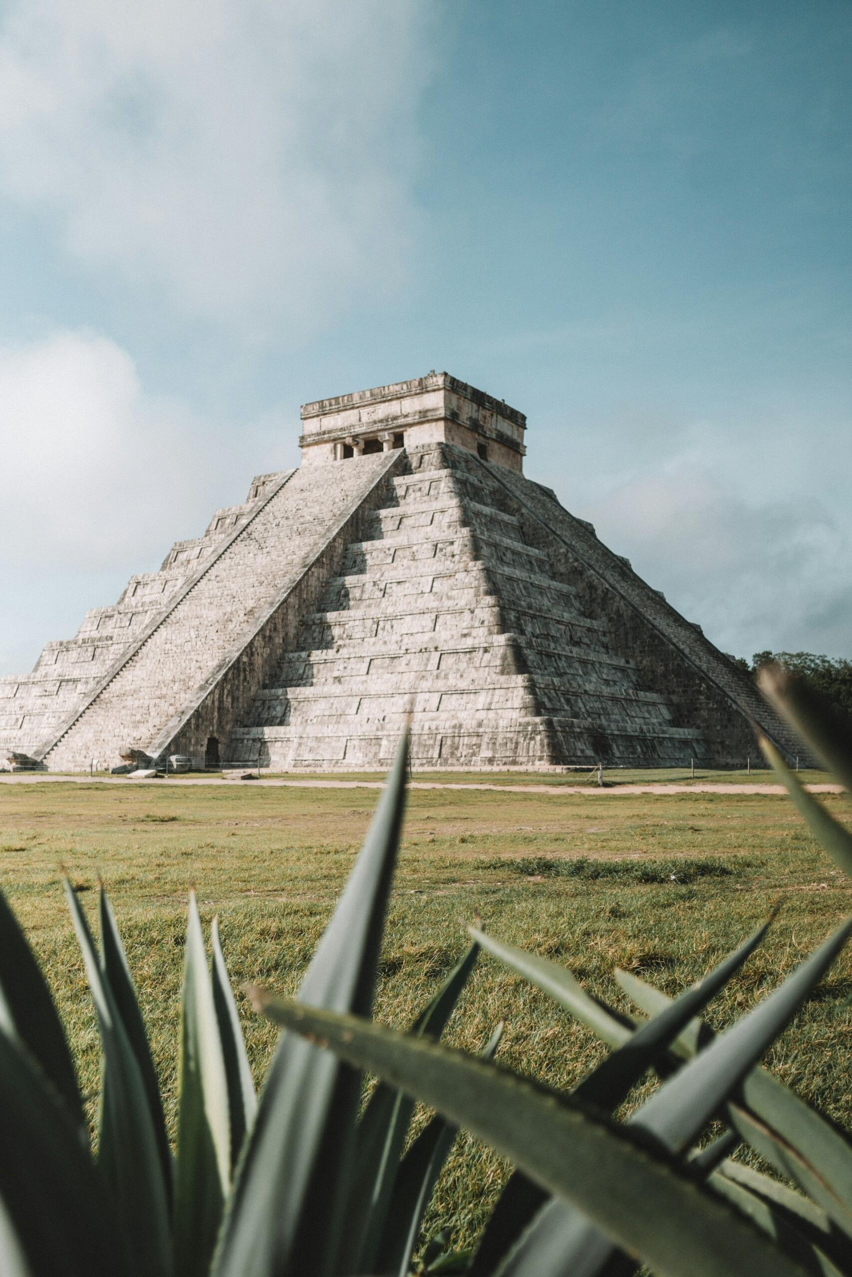 Una pirámide antigua bajo un cielo azul parcialmente nublado, rodeada de césped verde y vegetación en primer plano, lo que crea un ambiente histórico y sereno.