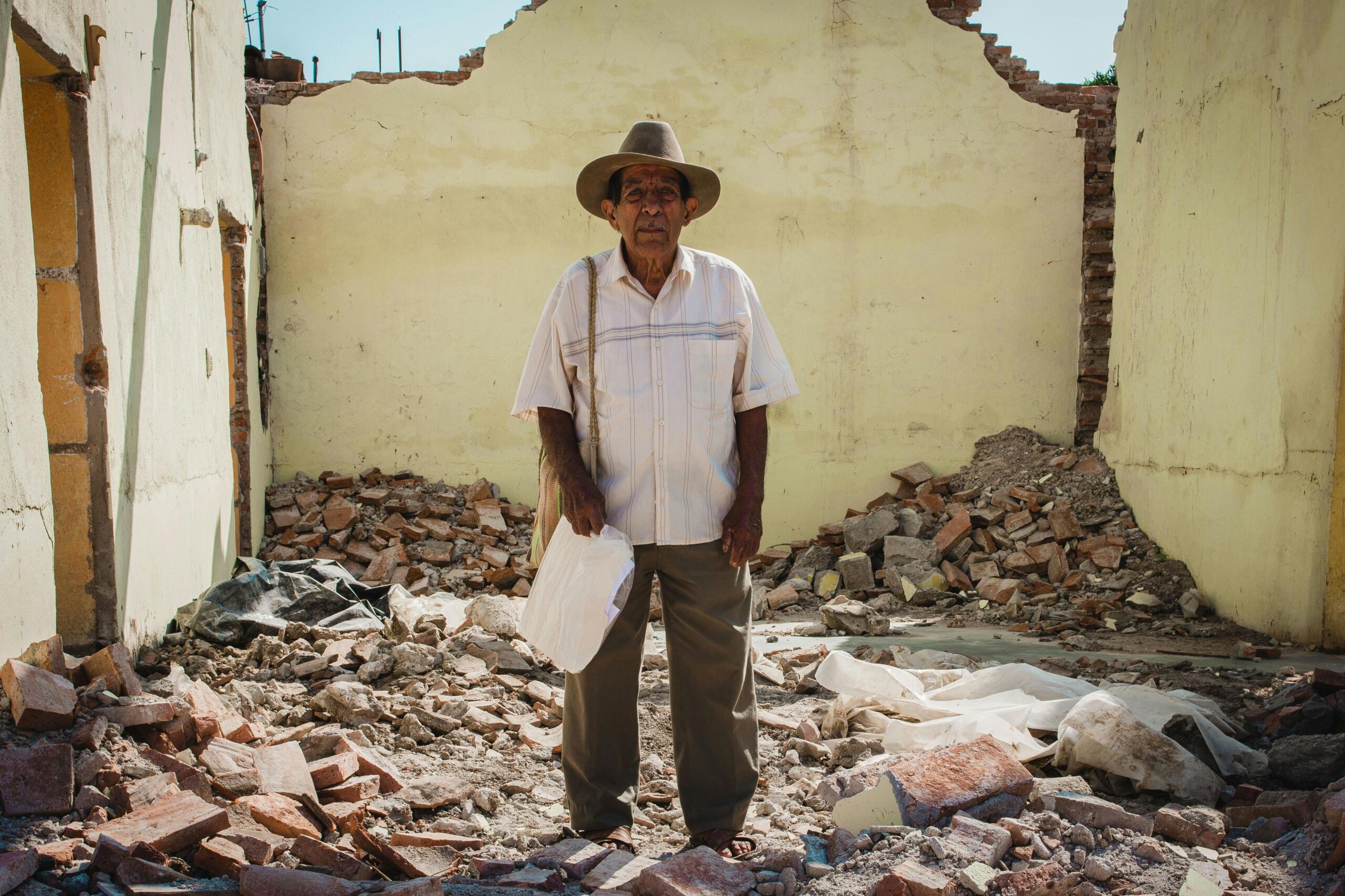 Un anciano con sombrero se encuentra de pie entre los escombros, en el interior de un edificio de paredes amarillas parcialmente derribado, durante el día.