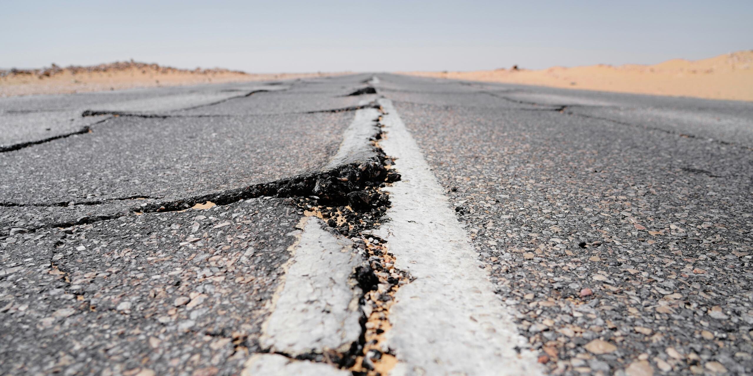 Primer plano de una carretera asfaltada agrietada y deteriorada, con una línea central blanca que atraviesa un paisaje desértico bajo un cielo despejado.