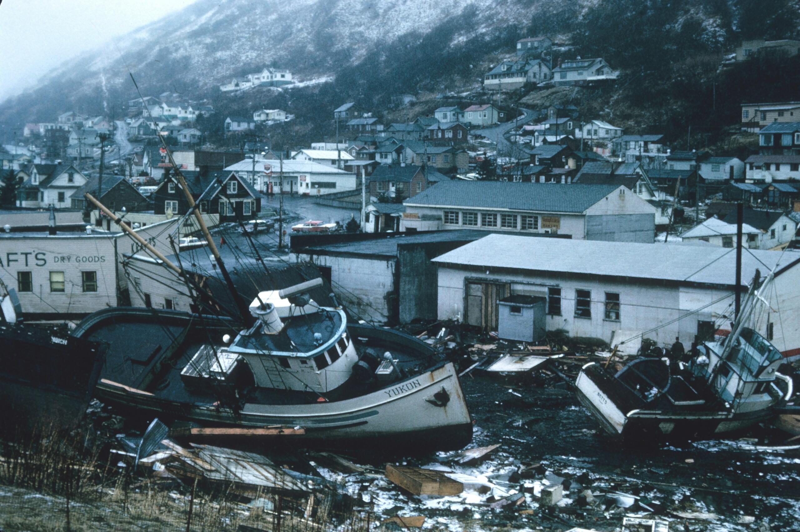 Dos barcos de pesca varados en un terreno lleno de escombros, cerca de unos edificios, en una localidad costera envuelta en niebla, con casas en la ladera de una colina.