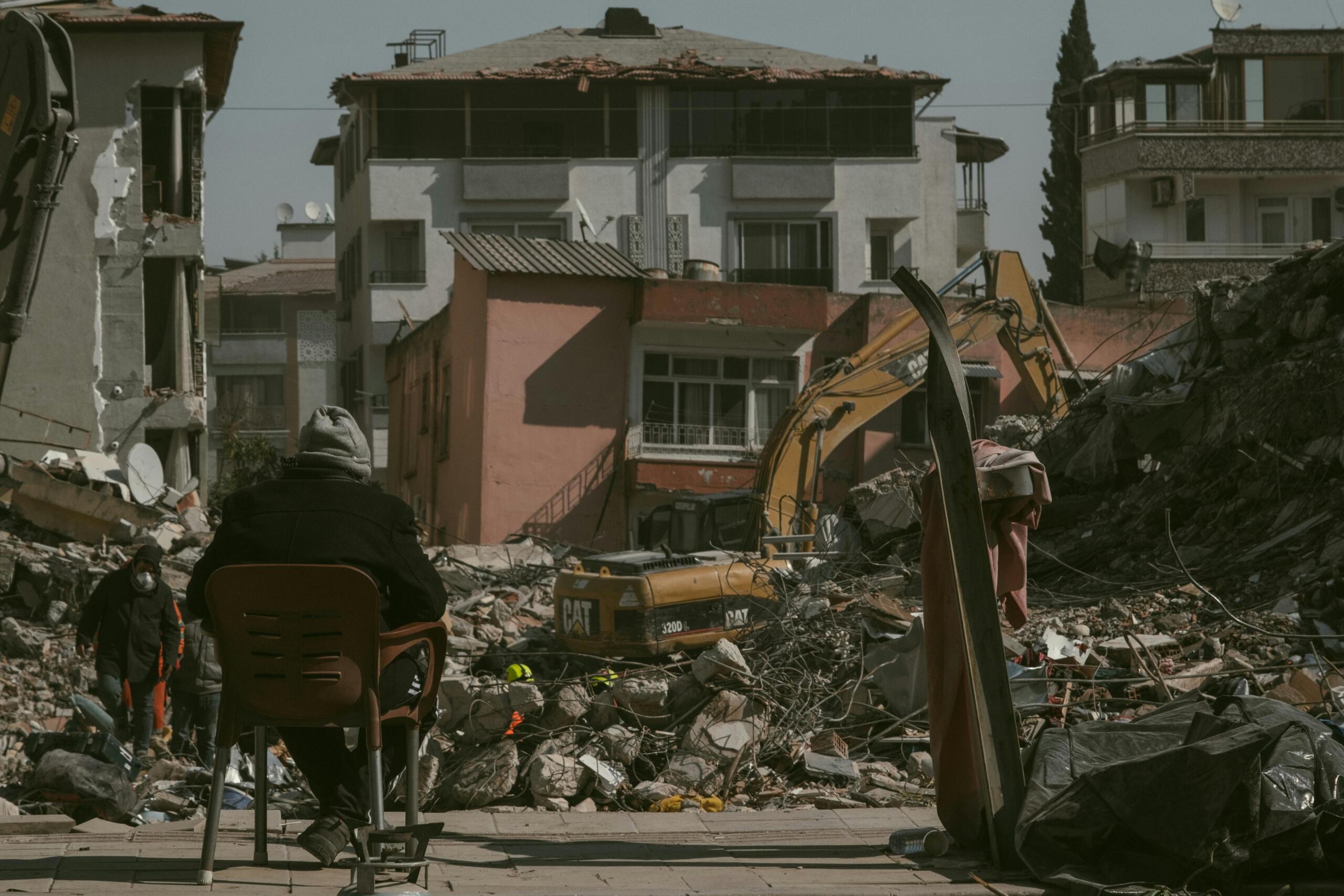Una persona sentada en una silla observa un lugar en obras de demolición con escombros, excavadoras y edificios dañados a la luz del día.