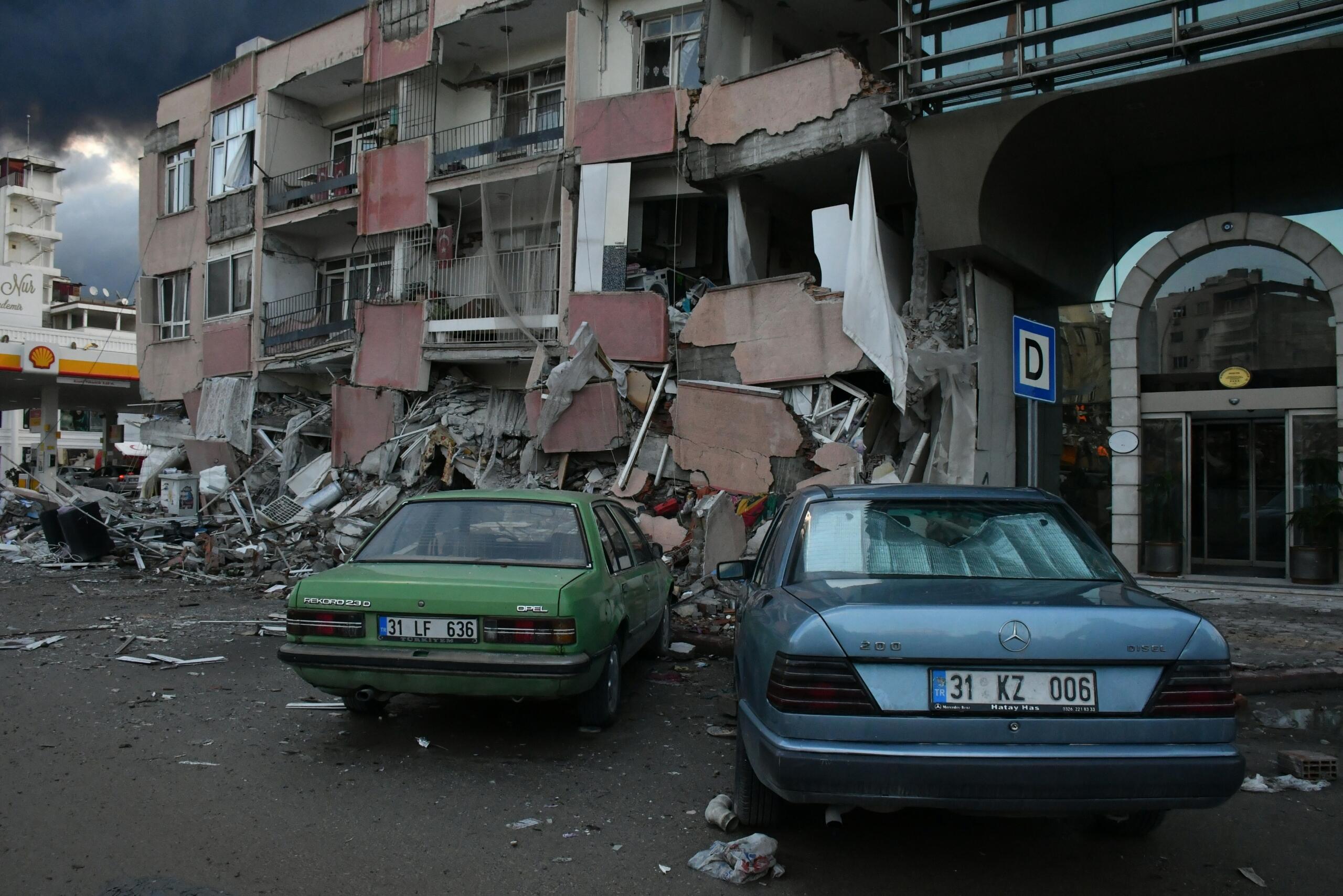 Edificio dañado con paredes derrumbadas y escombros; dos coches aparcados delante, bajo un cielo nublado y oscuro.