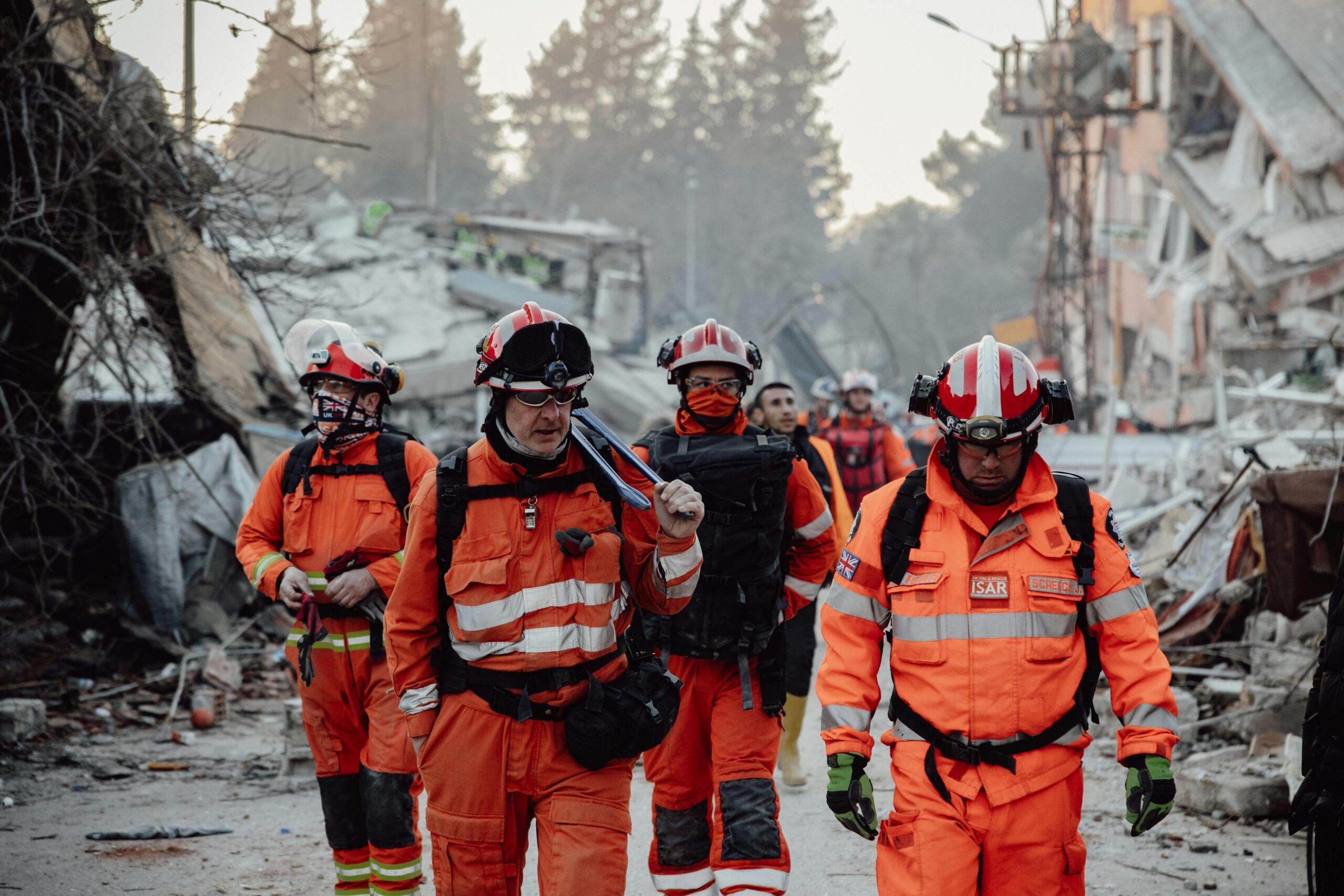Equipo de búsqueda y rescate con uniformes naranjas caminando entre los escombros de los edificios derrumbados tras una catástrofe.